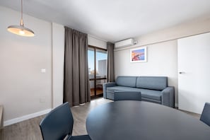 Una sala moderna con una mesa redonda, dos sillas, un sofá gris, una lámpara colgante y un cerramiento de vidrio corredero que da a un balcón.