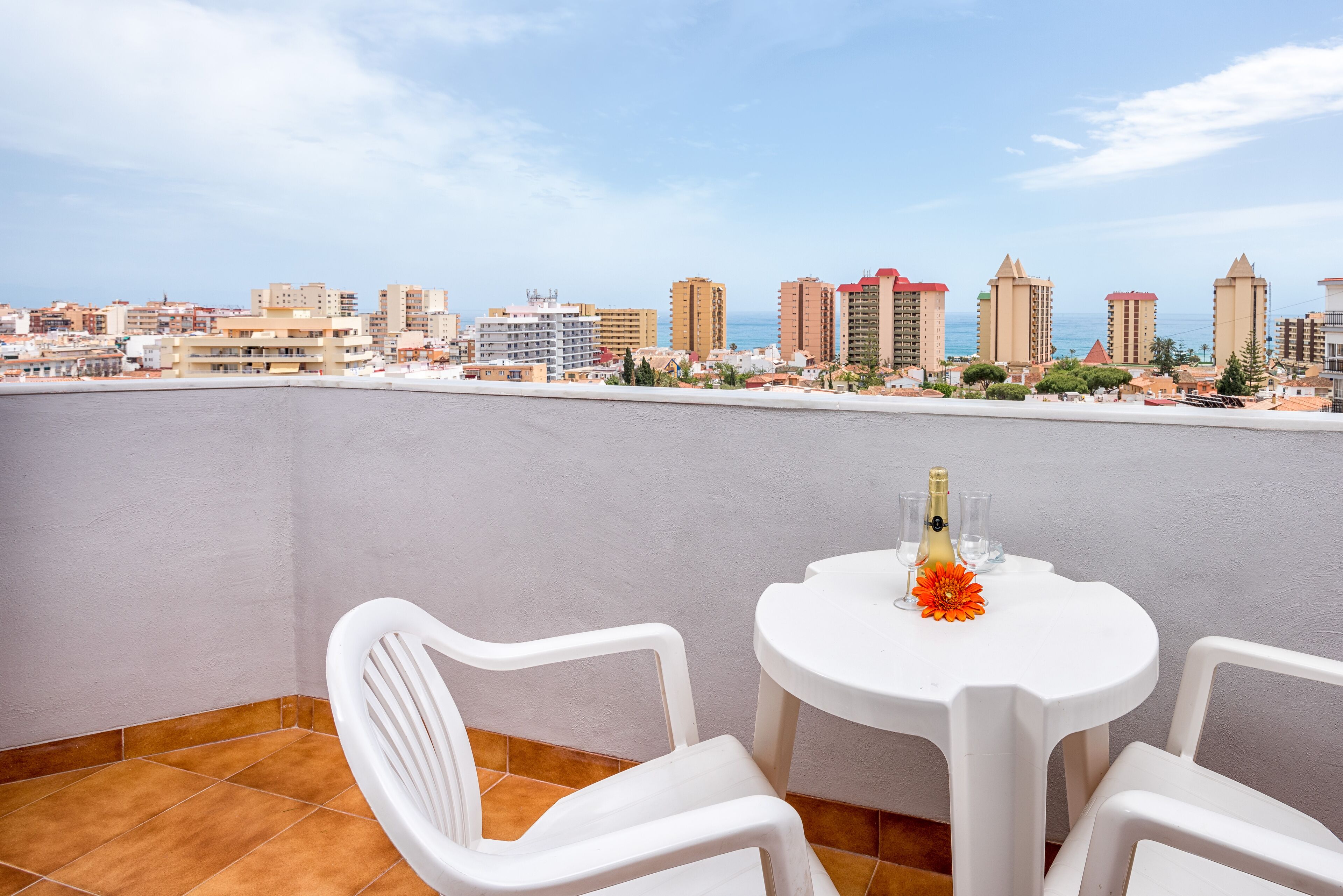Una terraza con sillas y una mesa de plástico blanco, con vistas a un paisaje urbano con diversos edificios y un cielo despejado.