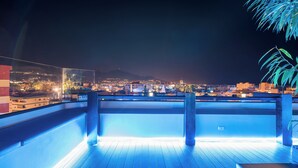 Vista nocturna desde un balcón con vistas a un paisaje urbano de edificios iluminados y una montaña distante.