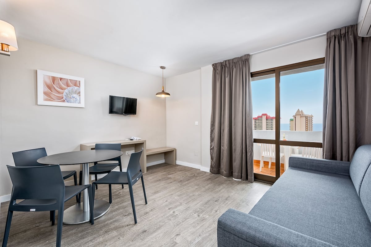 Ett modernt vardagsrum med en grå soffa, ett matbord med stolar, en platt-TV och ett stort fönster med gardiner.