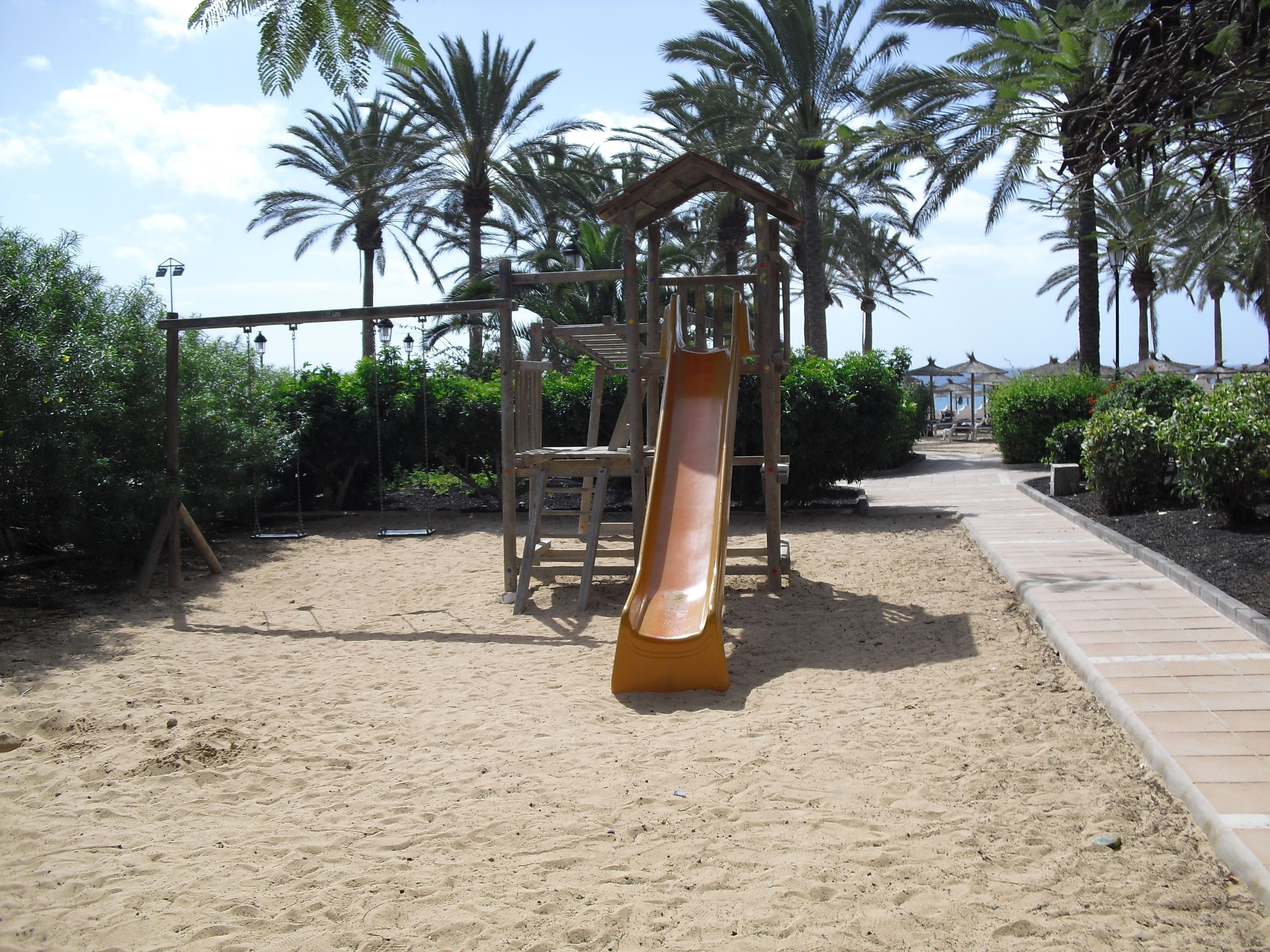 A playground with a slide, swings, and a sandy ground surrounded by palm trees.