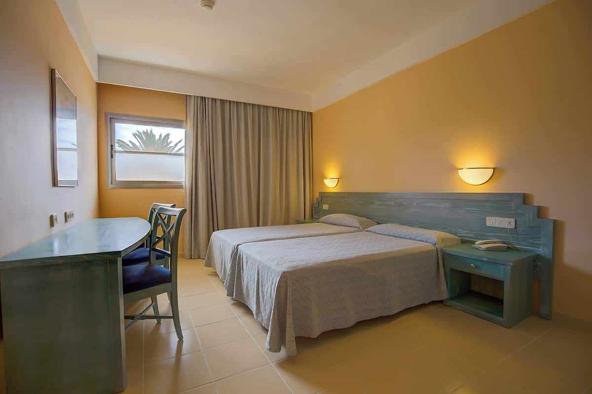 A hotel room with a bed, a desk, a chair, two bedside tables, and a window with curtains.