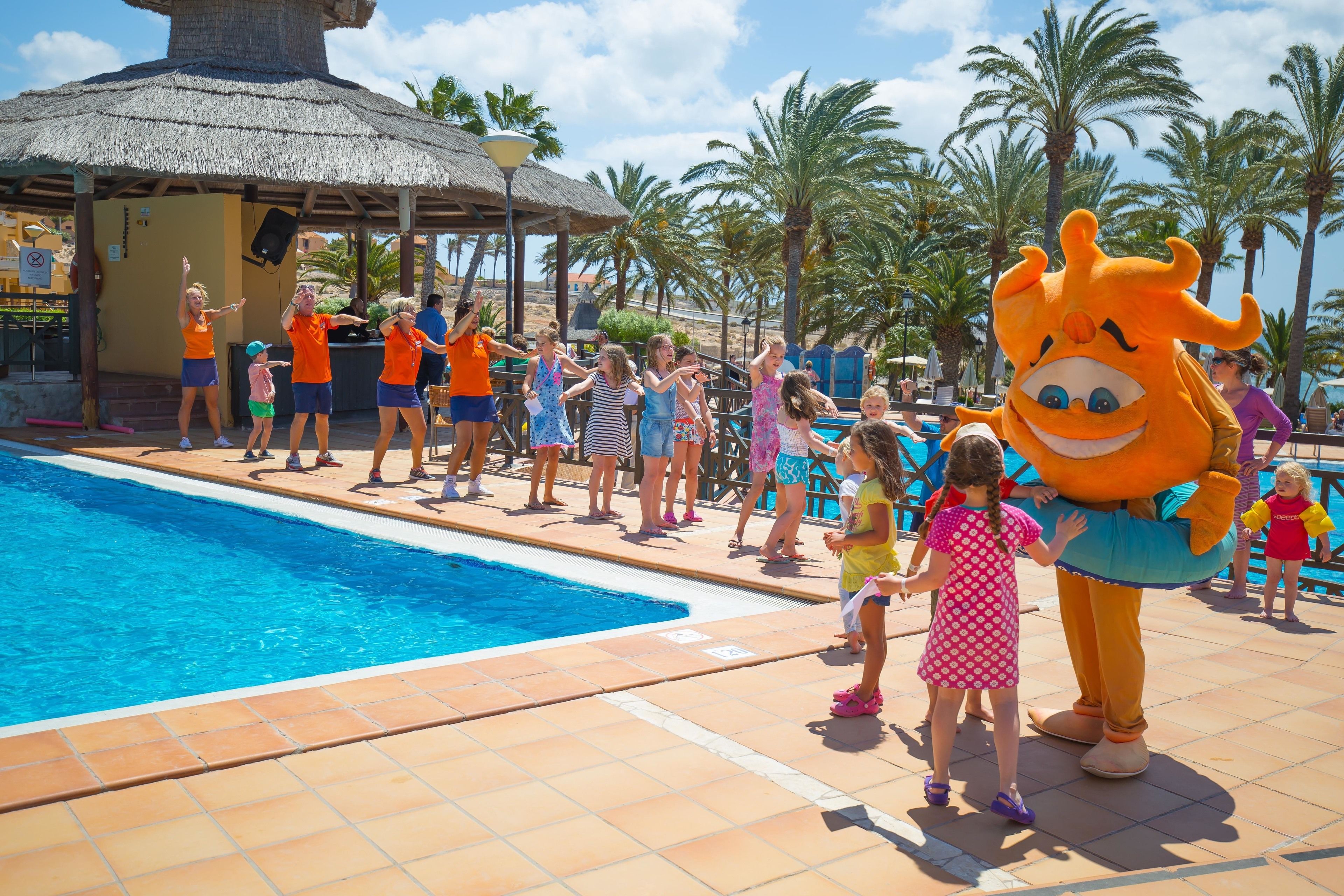 Un área de piscina con una estructura de techo de paja, palmeras y un personaje mascota.