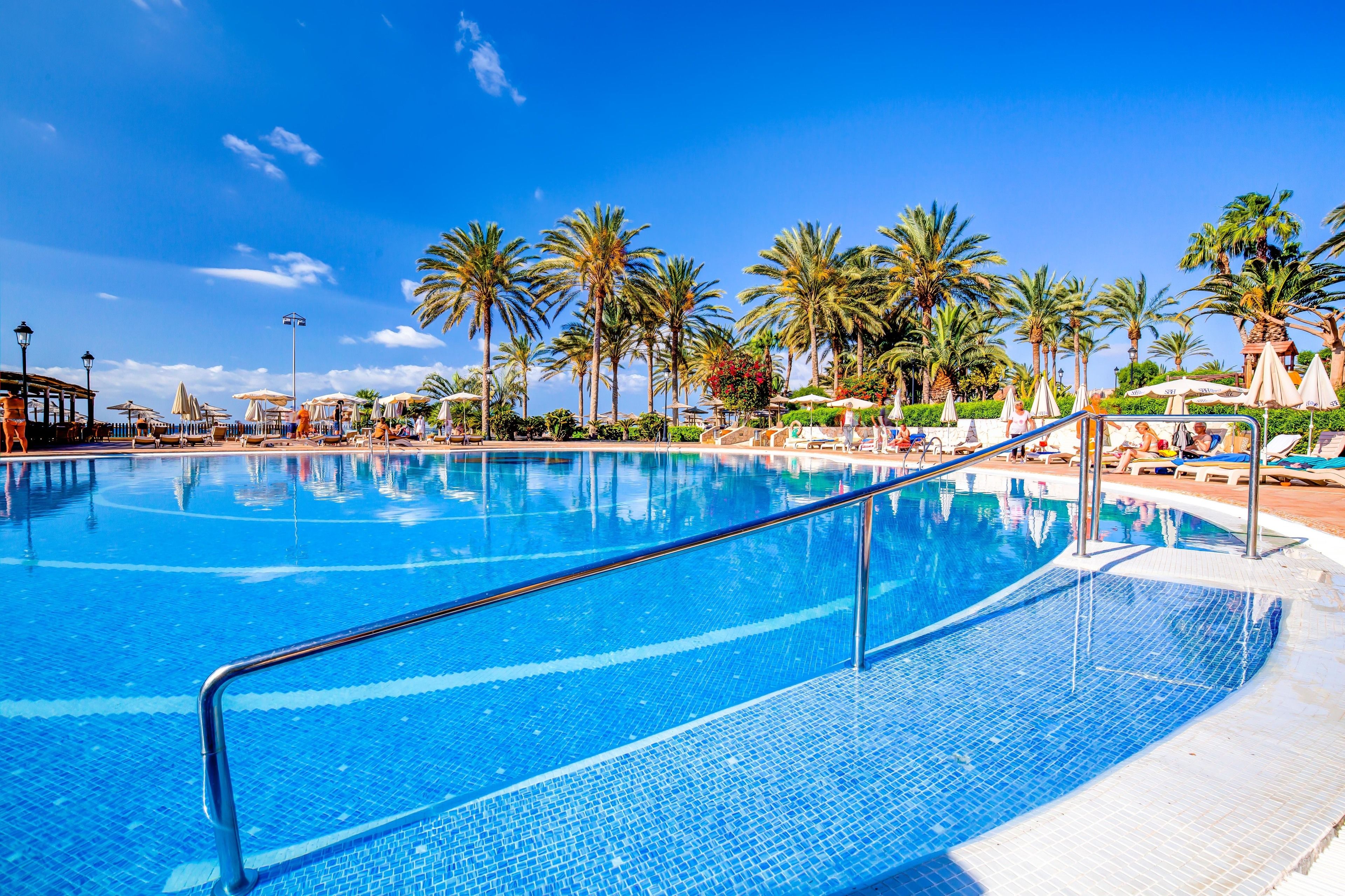 A large, clear blue pool with a metal railing, surrounded by palm trees and sun loungers under umbrellas.