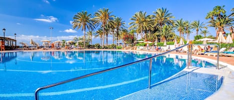 A large, clear blue pool with a metal railing, surrounded by palm trees and sun loungers under umbrellas.