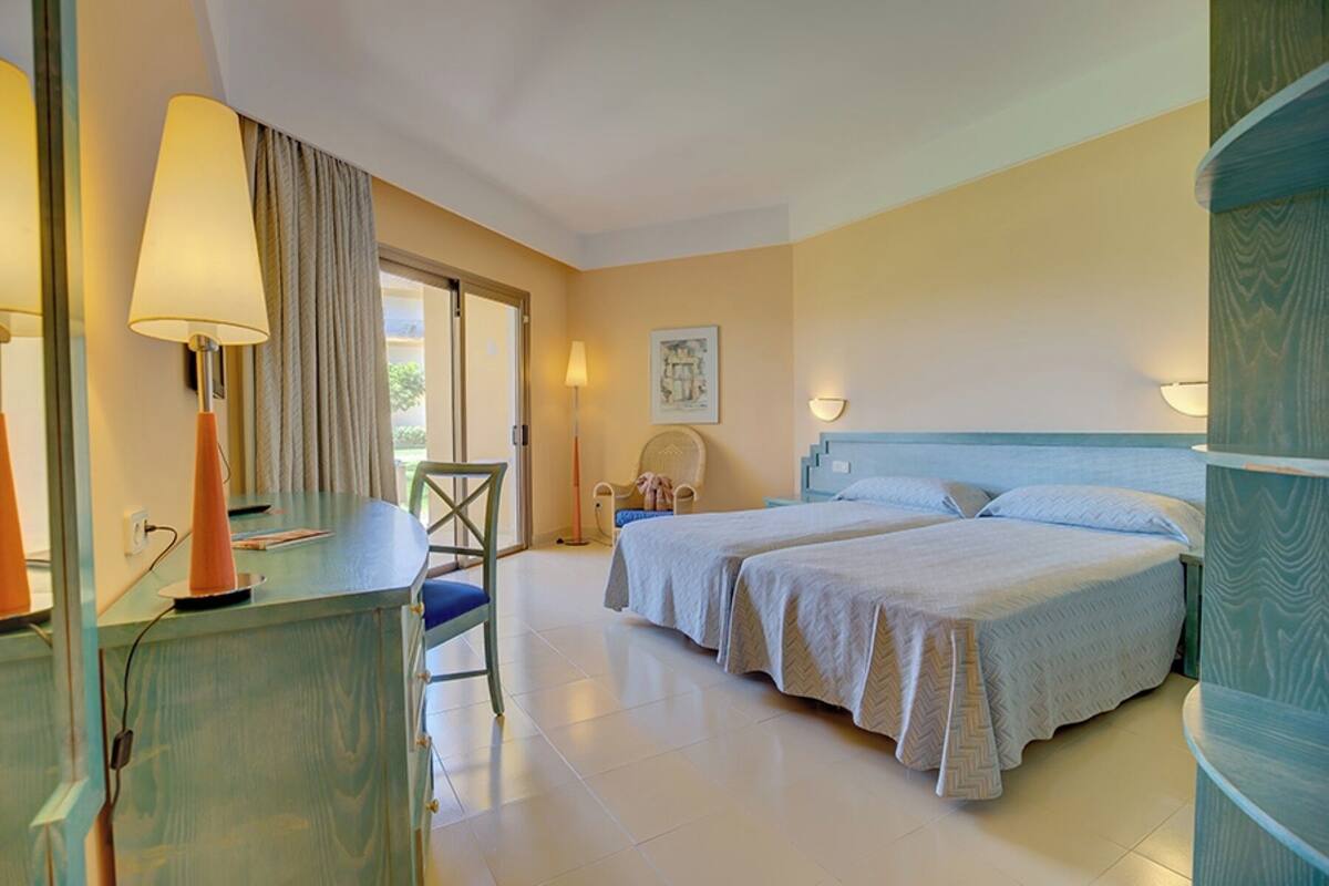 A hotel room with two beds, a desk with a lamp, a chair, and a window with curtains.