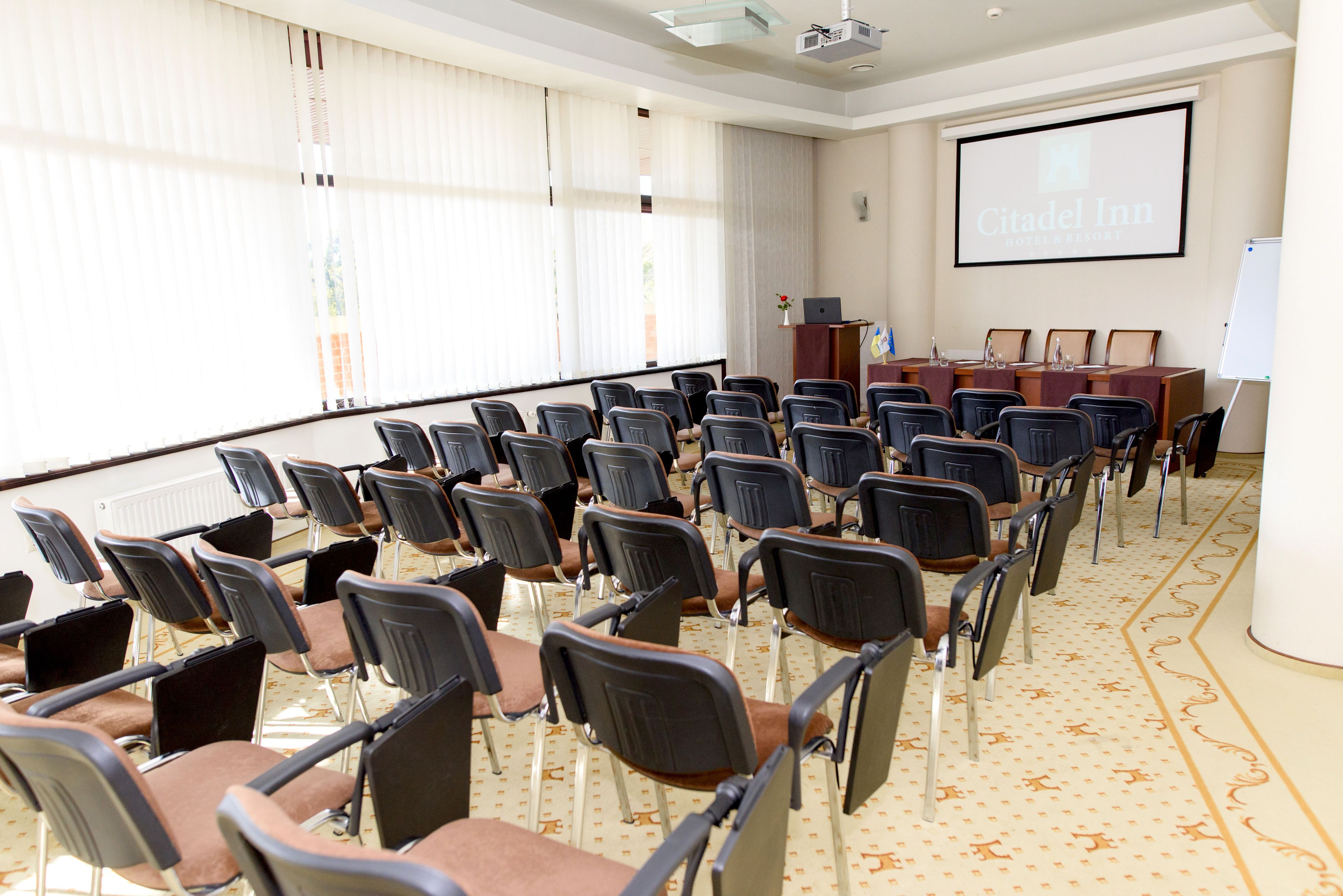 Ein Konferenzraum mit Reihen von Stühlen, einem Rednerpult und einem Bildschirm, der 'Citadel Inn Hotel & Resort' anzeigt.