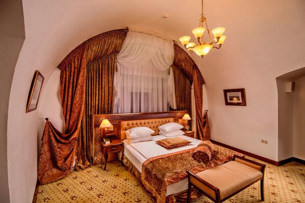 A bedroom with a large bed, a nightstand, a chair, a chandelier, and curtains.