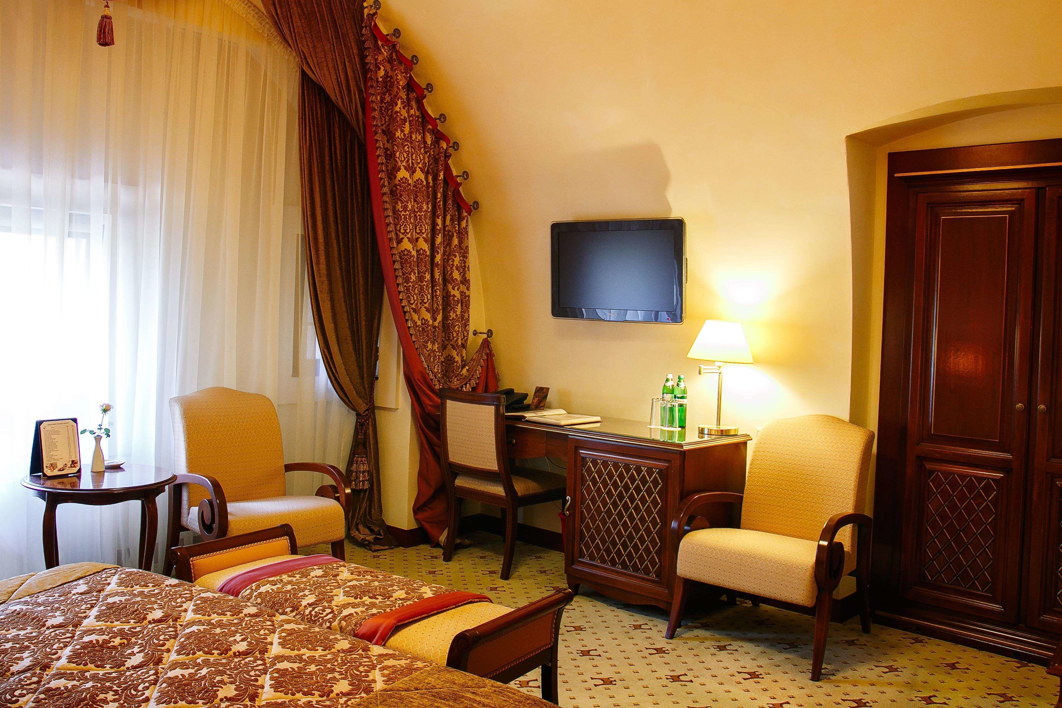 A hotel room with a bed, two armchairs, a desk, a television, and a wardrobe.