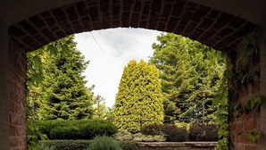 Een gebogen bakstenen constructie omlijst een tuin met hagen, een hoge boom en roze bloemen.