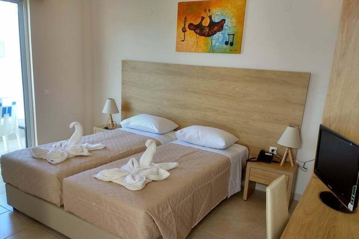 A hotel room with two beds, a wooden headboard, a painting above the beds, a desk with a computer, and a TV on the right.