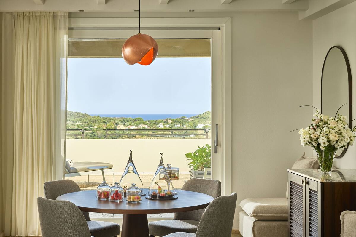 Een eetruimte met een ronde tafel, stoelen en uitzicht op de zee door een groot raam.