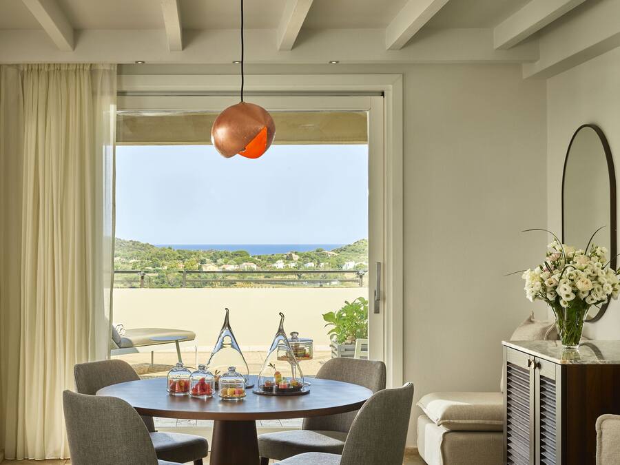 A dining area with a round table, chairs, and a view of the sea through a large window.