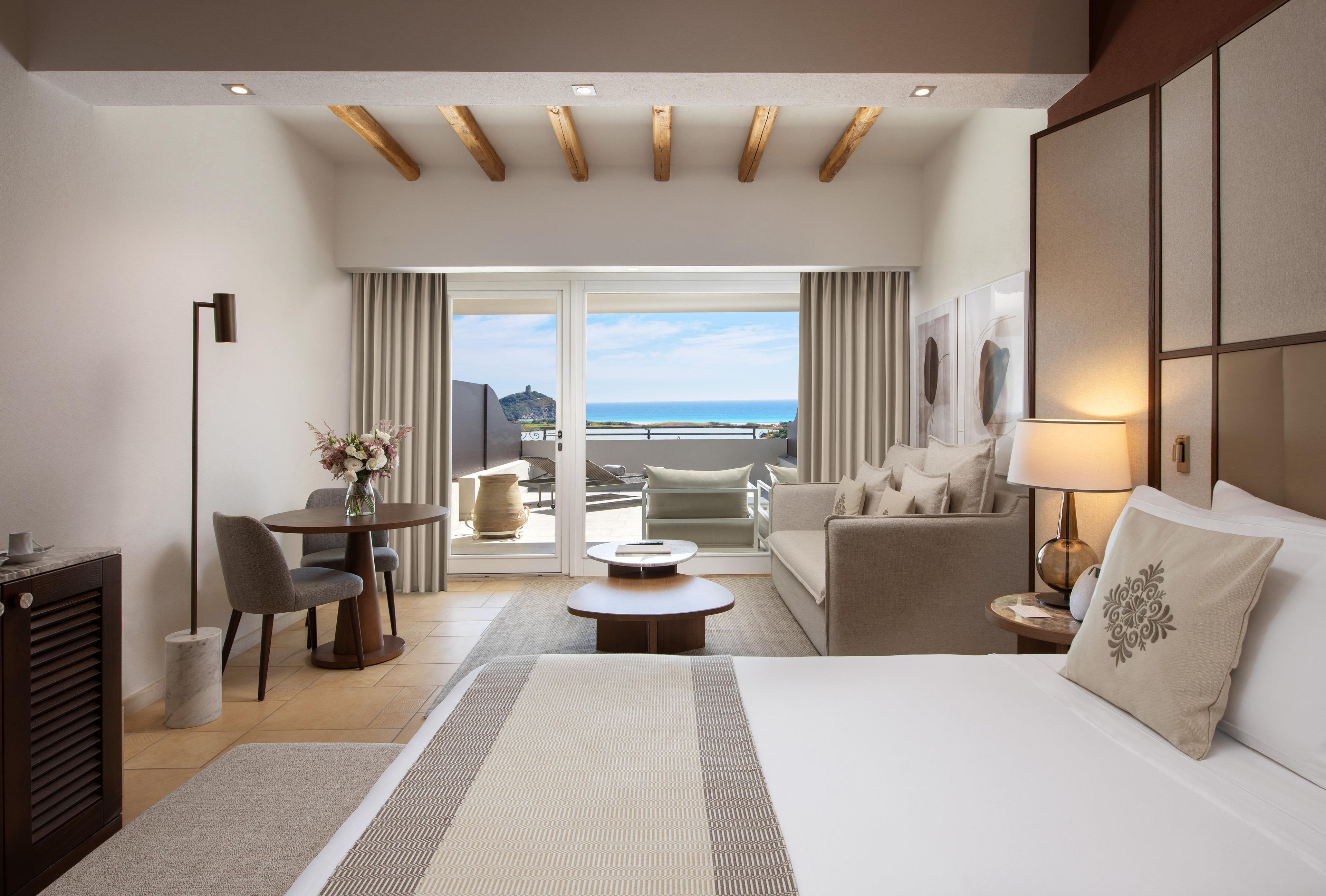 Ein Hotelzimmer mit Bett, einer Couch, einem Esstisch mit Stühlen, einem Schreibtisch und Blick auf den Ozean durch große Fenster.