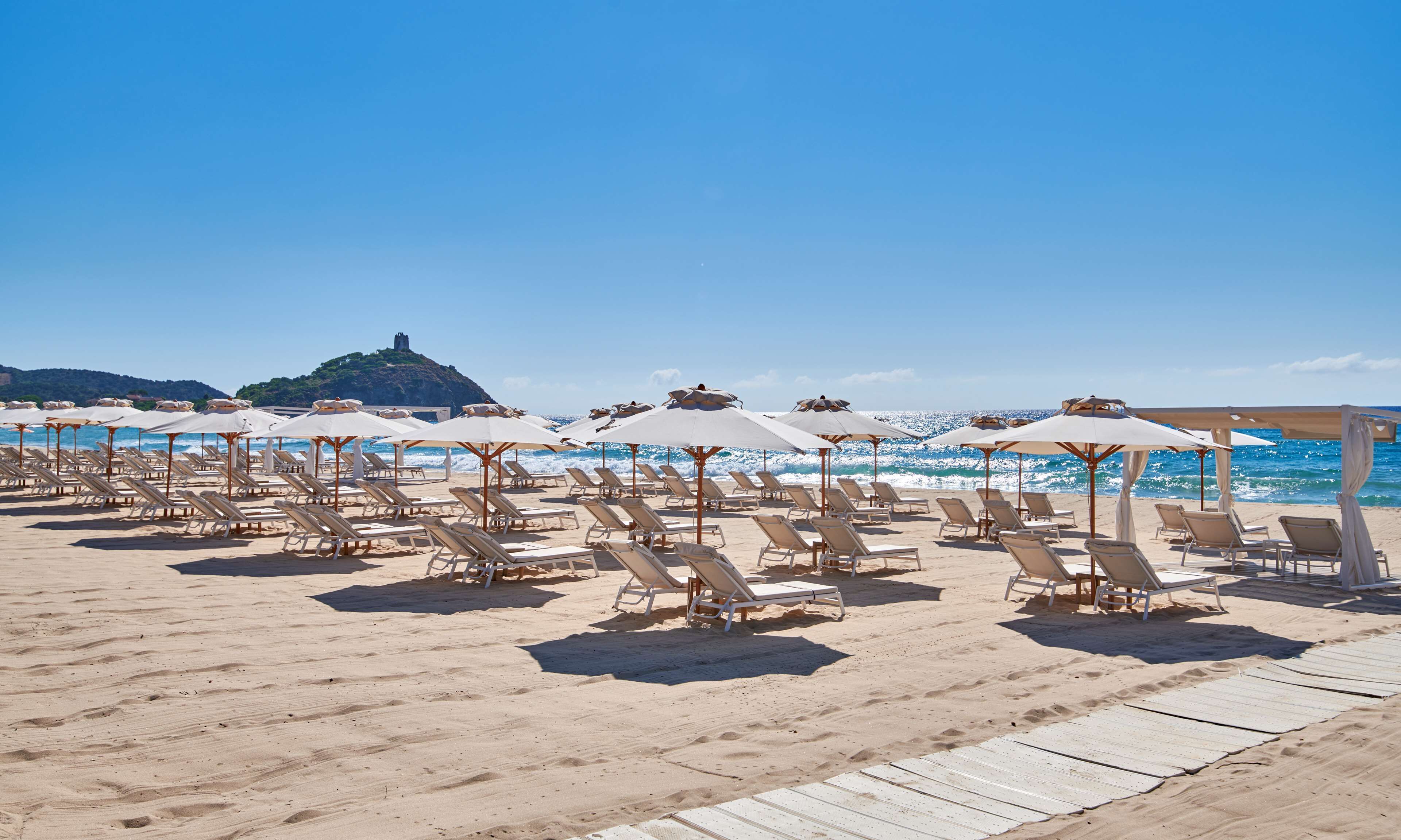 Una playa con filas de tumbonas y sombrillas, una terraza de madera y un castillo en la colina al fondo.