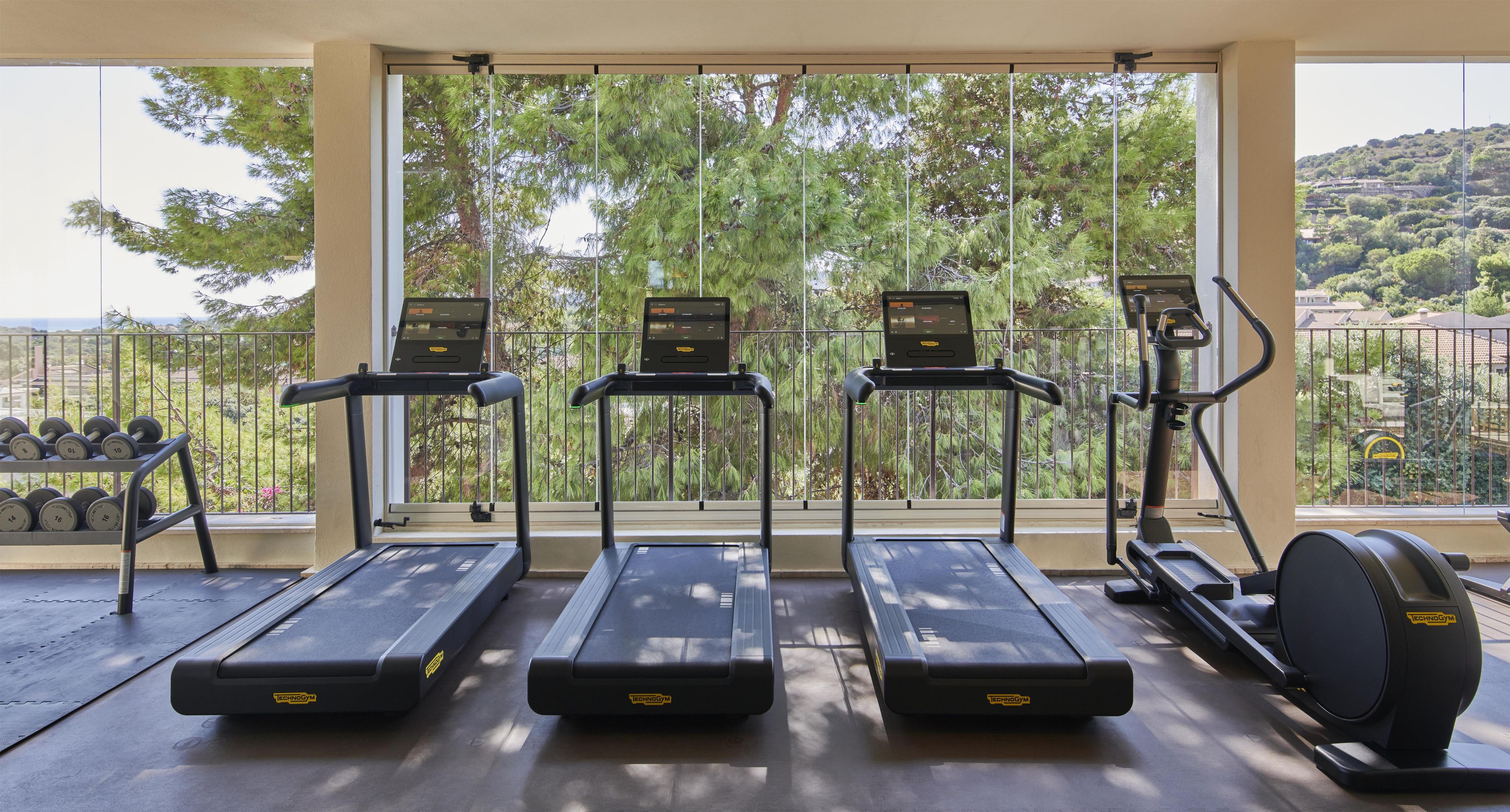 Un gimnasio con cintas de correr, mancuernas y una máquina elíptica, con vistas a árboles y un paisaje montañoso.