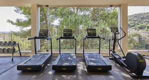 Une salle de sport équipée de tapis de course, d’haltères et d’un appareil elliptique, avec vue sur des arbres et un paysage vallonné.