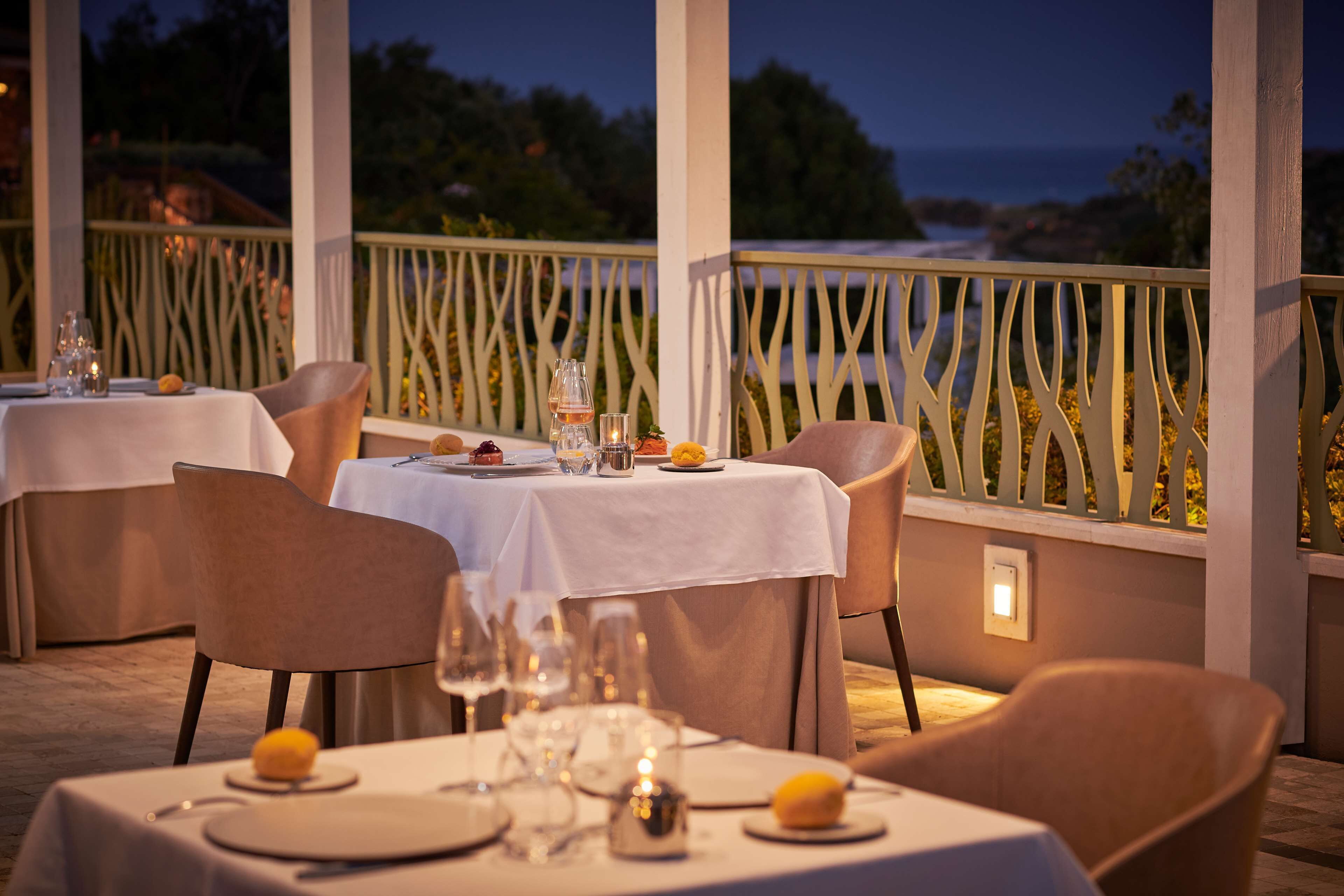 An outdoor dining area with tables set for two, featuring white tablecloths and candle centerpieces.