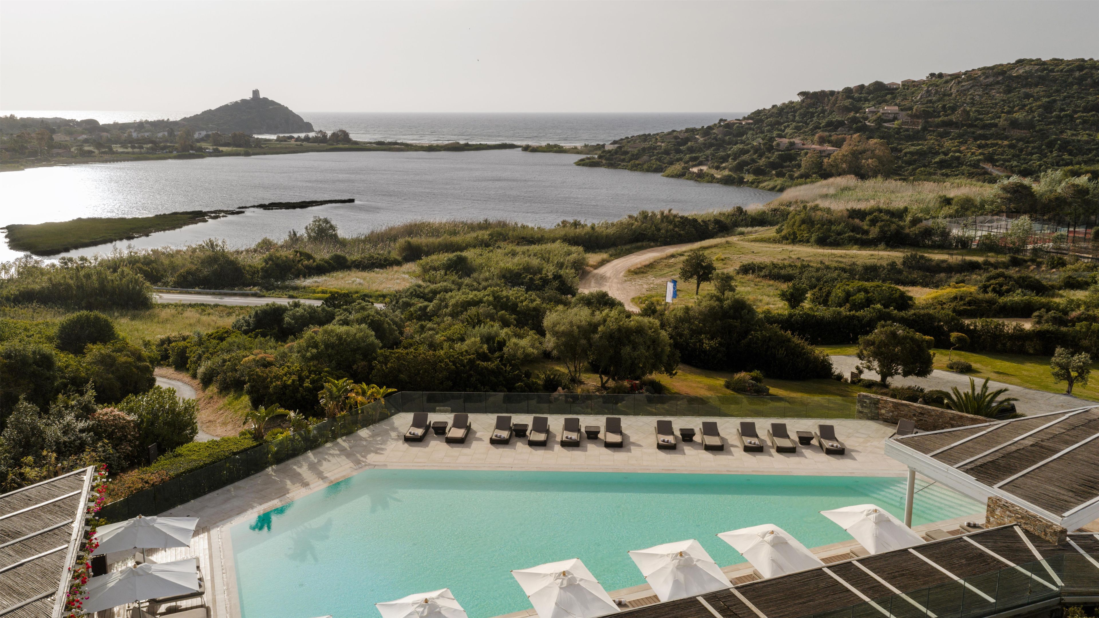 A pool with sun loungers and umbrellas, surrounded by greenery and overlooking a body of water.