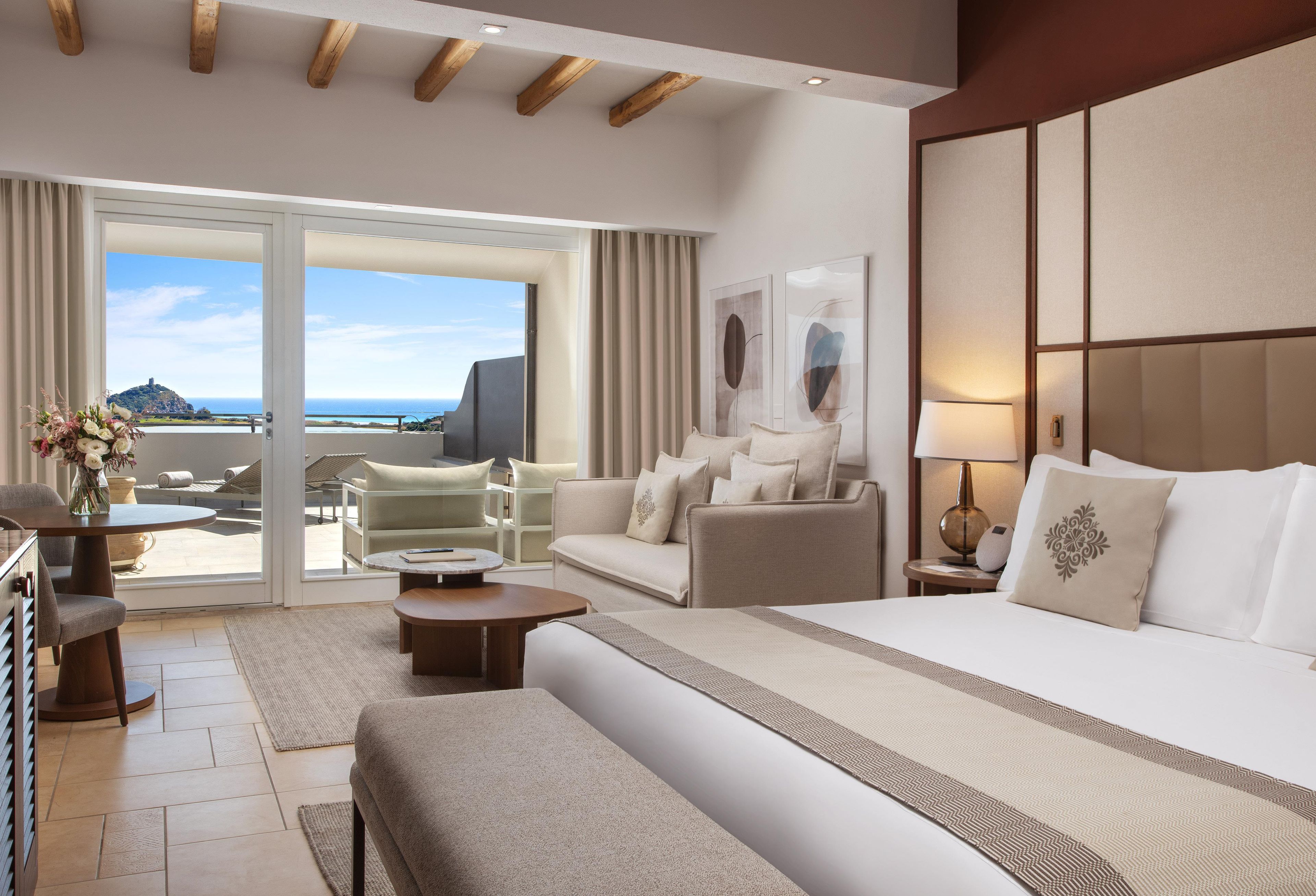 A hotel room with a bed, a sofa, a chair, a table, and a view of the sea through a sliding door.