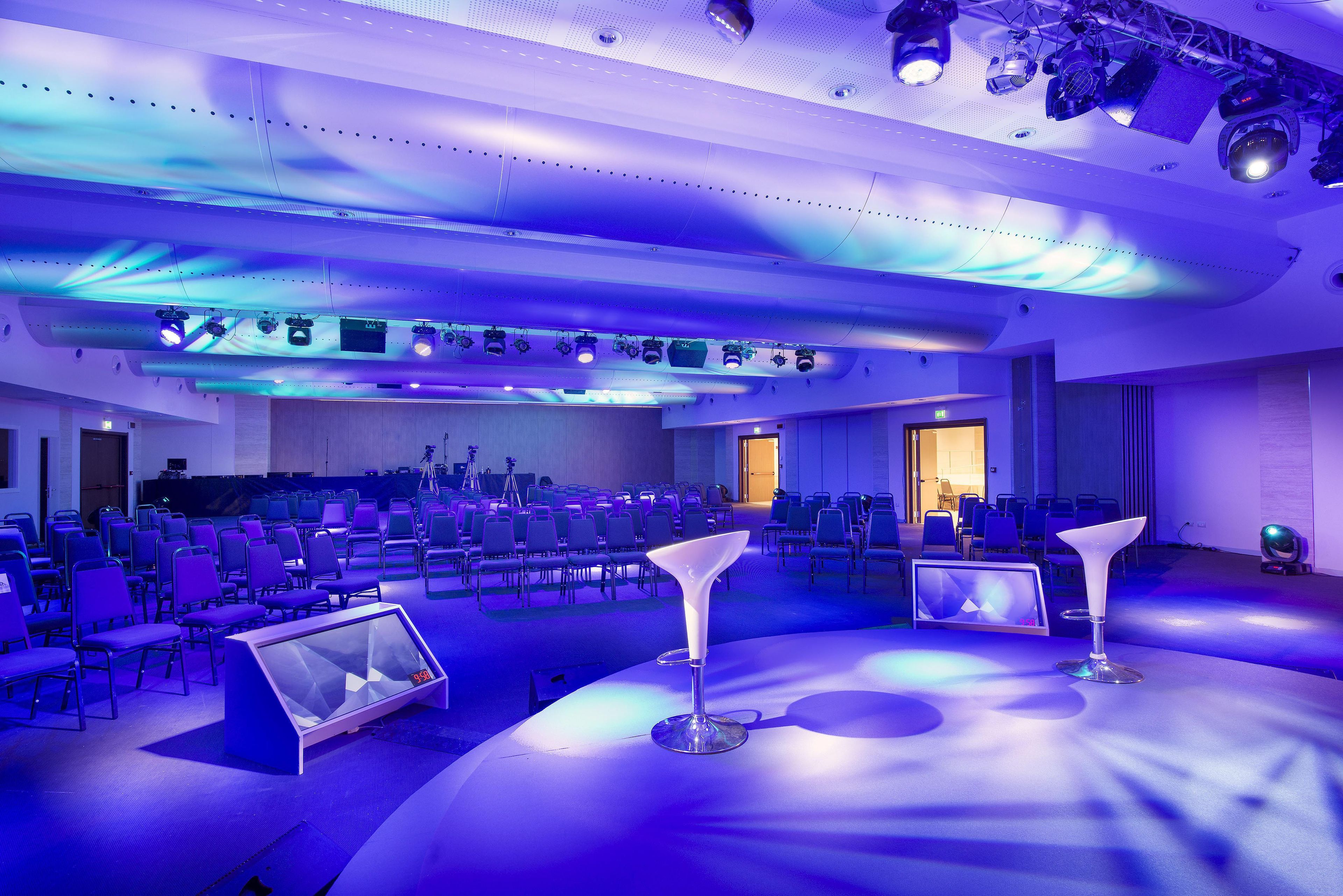 A conference room with rows of chairs, a stage with a podium, and ceiling-mounted lighting.