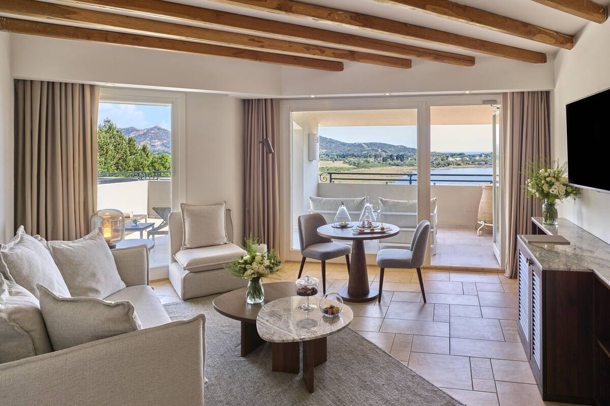 Een ruime woonkamer met een bank, salontafel en stoelen, een balkon met uitzicht op de bergen, en een aan de muur bevestigde televisie.