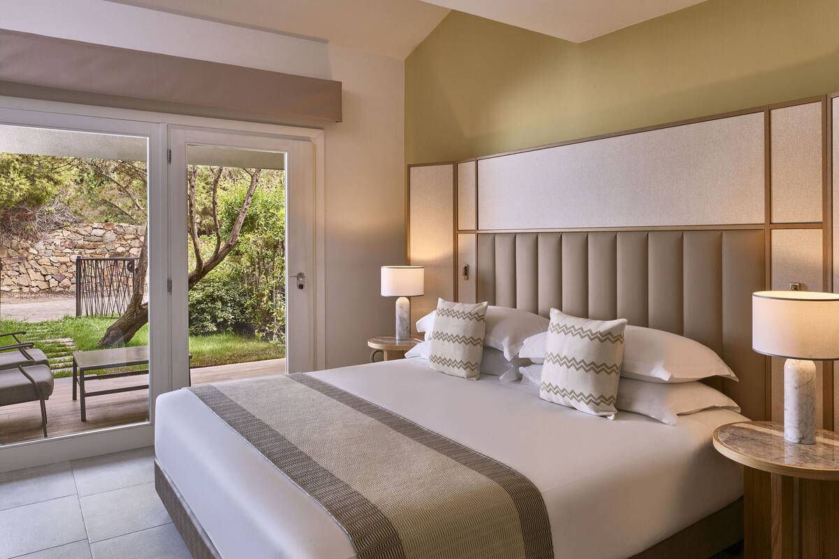 Een hotelkamer met een groot bed, twee bedlampjes, uitzicht op de omgeving en een stenen muur.