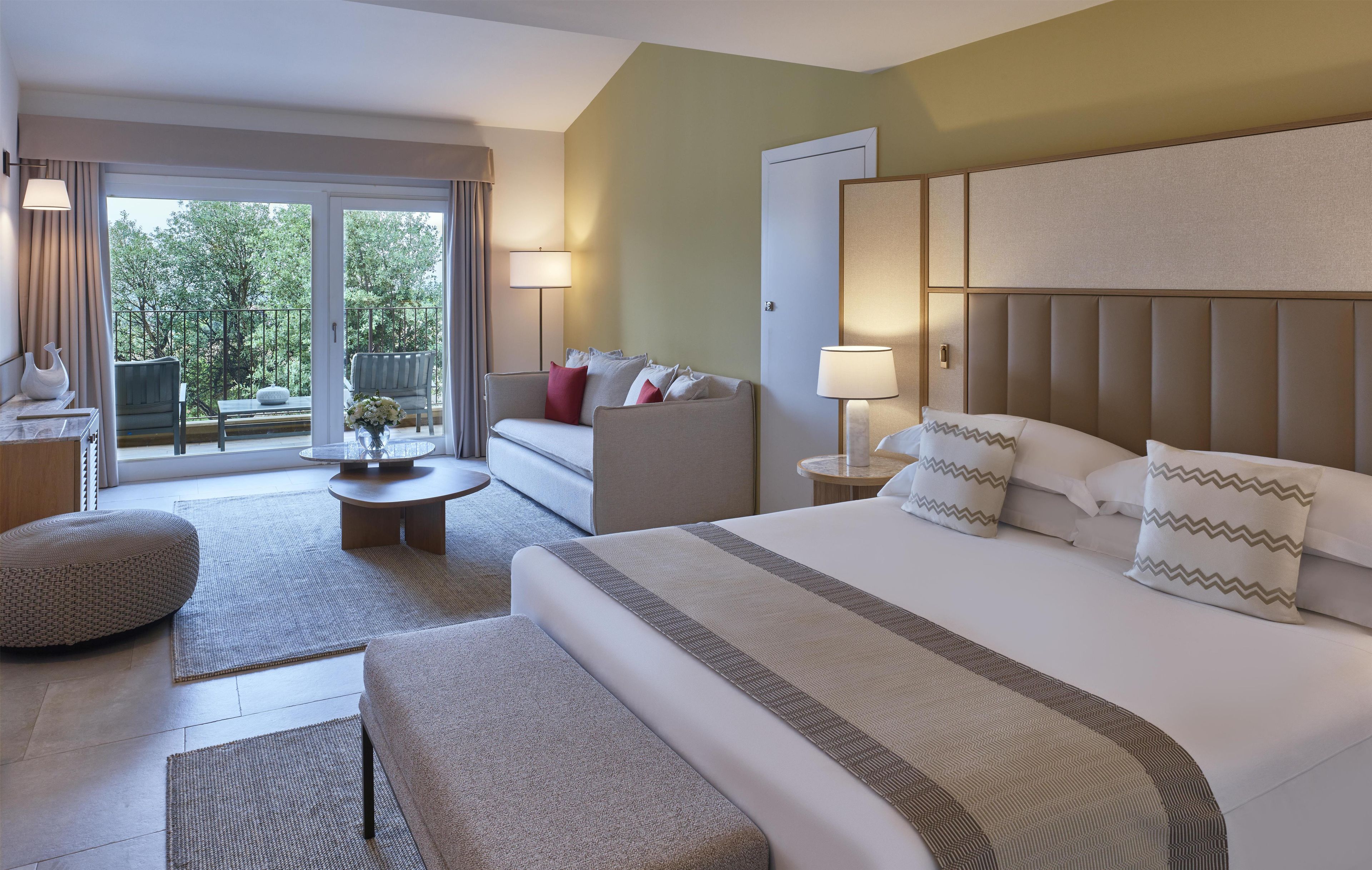 Habitación de hotel con una cama grande, un sofá, una mesita y un balcón con vistas a árboles.