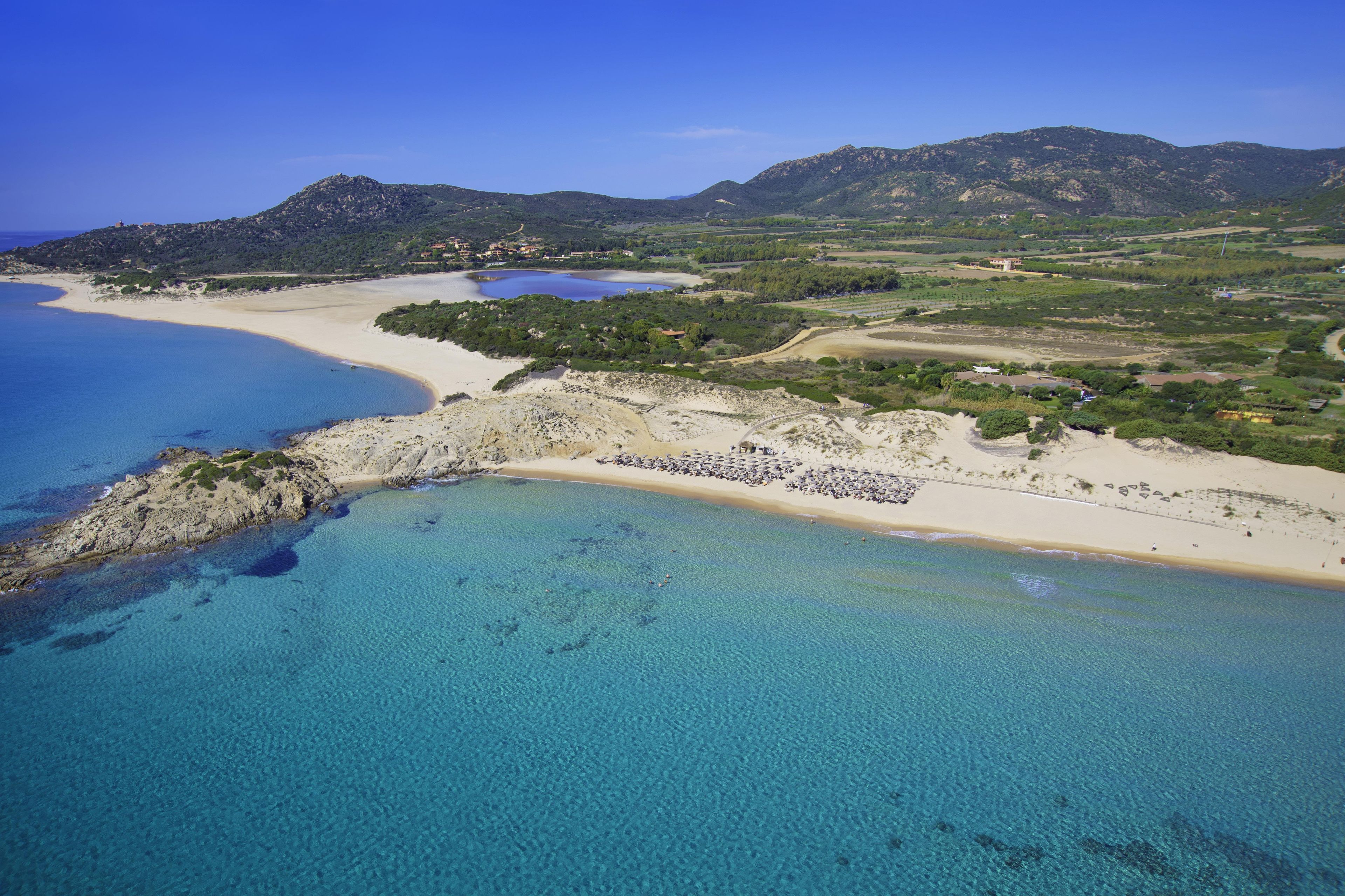 Vista aérea de una zona costera con una playa de arena, aguas azules cristalinas y colinas verdes al fondo.