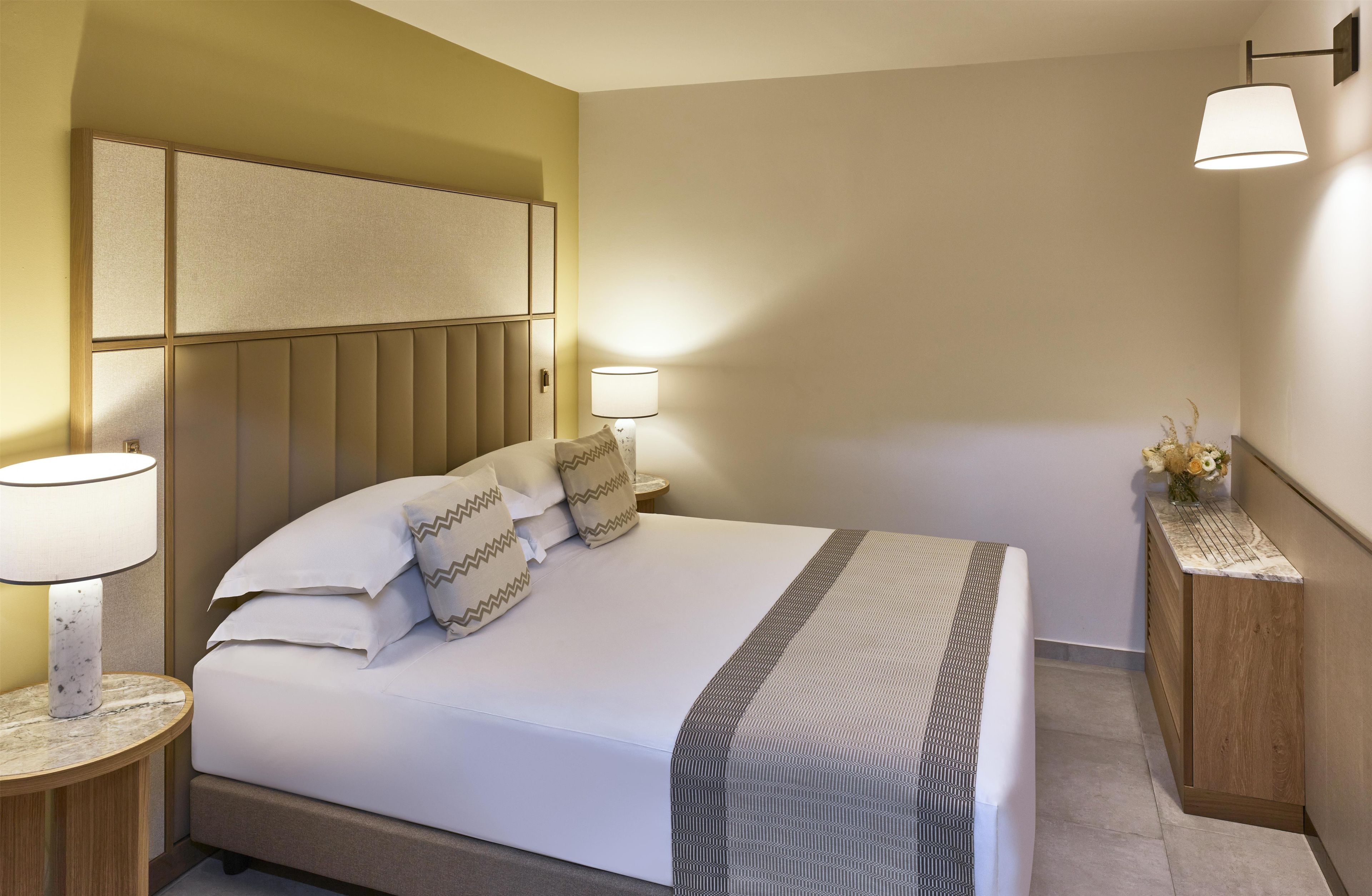 Een hotelkamer met een groot bed, twee bedlampen, een hoofdeinde en een klein nachtkastje met een bloemenvaas.