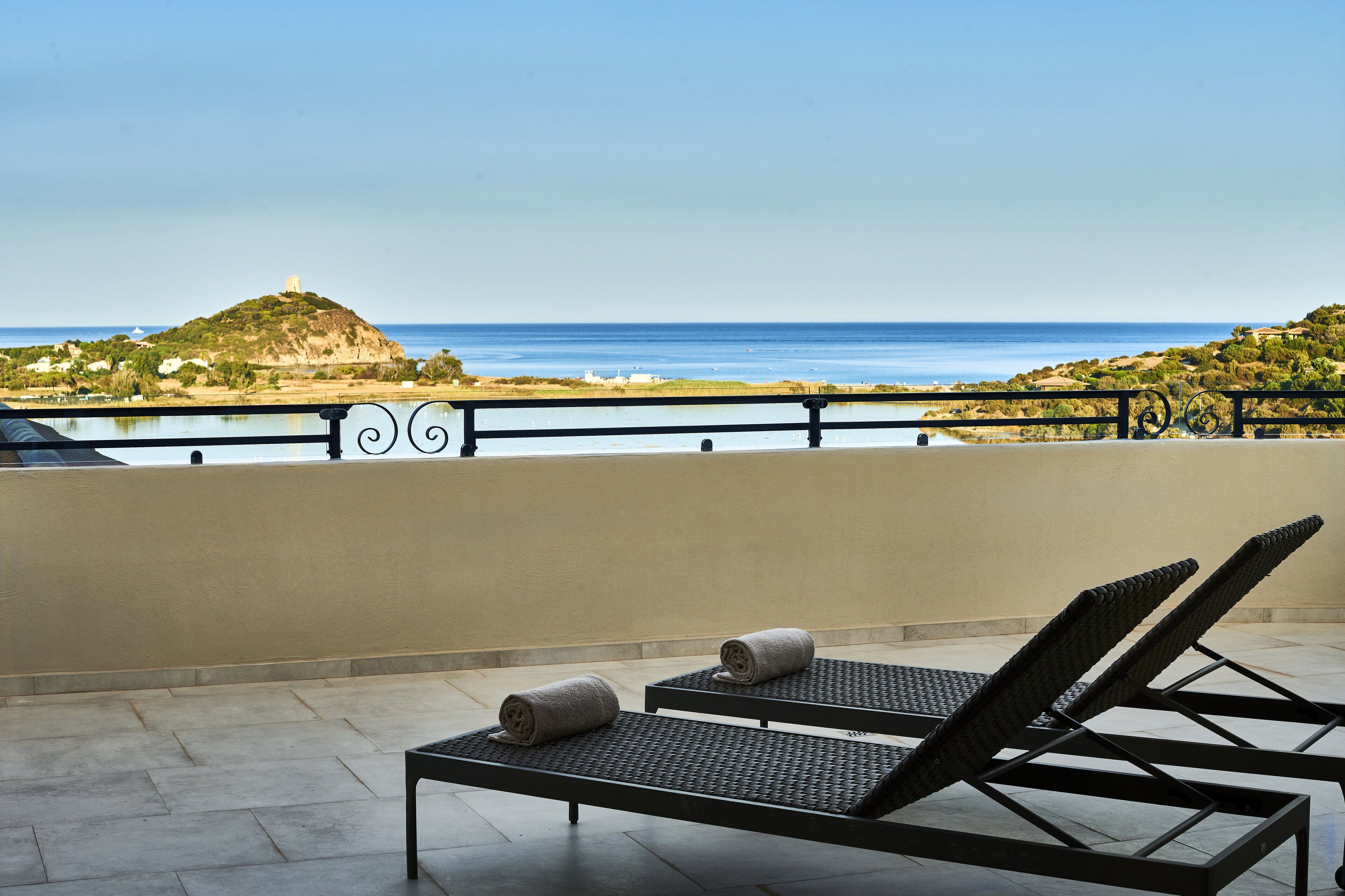 Ein Balkon mit zwei Lounge-Stühlen und einem Tisch, mit Blick auf eine Küstenlandschaft mit Hügel und Meer.