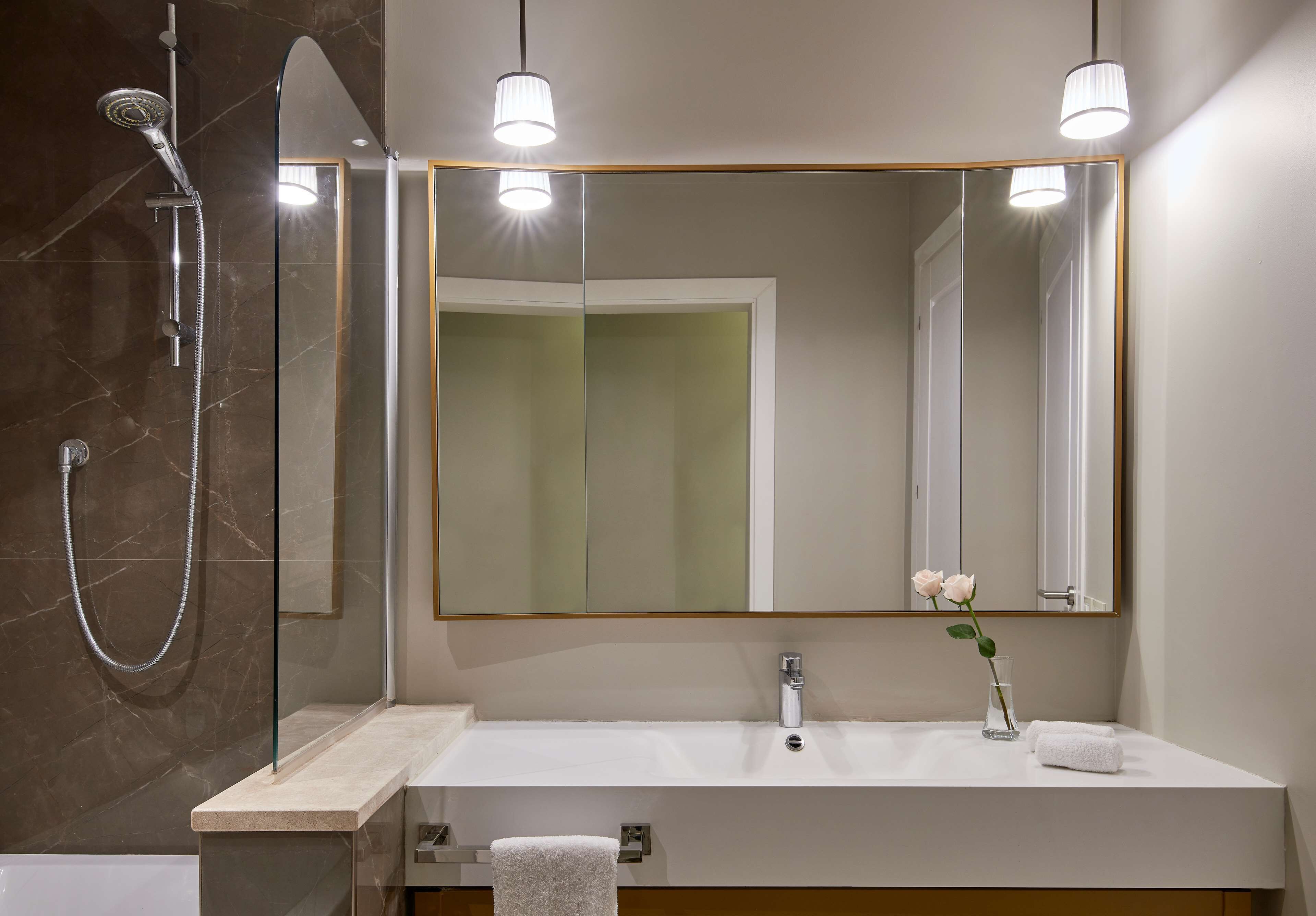 Un baño moderno con cabina de ducha de vidrio, un espejo grande, un lavabo blanco y un florero con una flor.