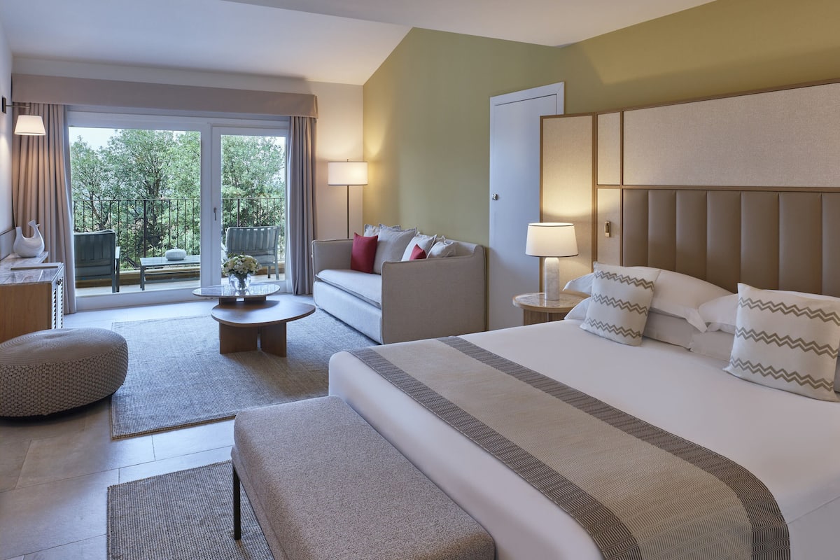 Een hotelkamer met een groot bed, een bank, een klein tafeltje en een balkon met uitzicht op bomen.