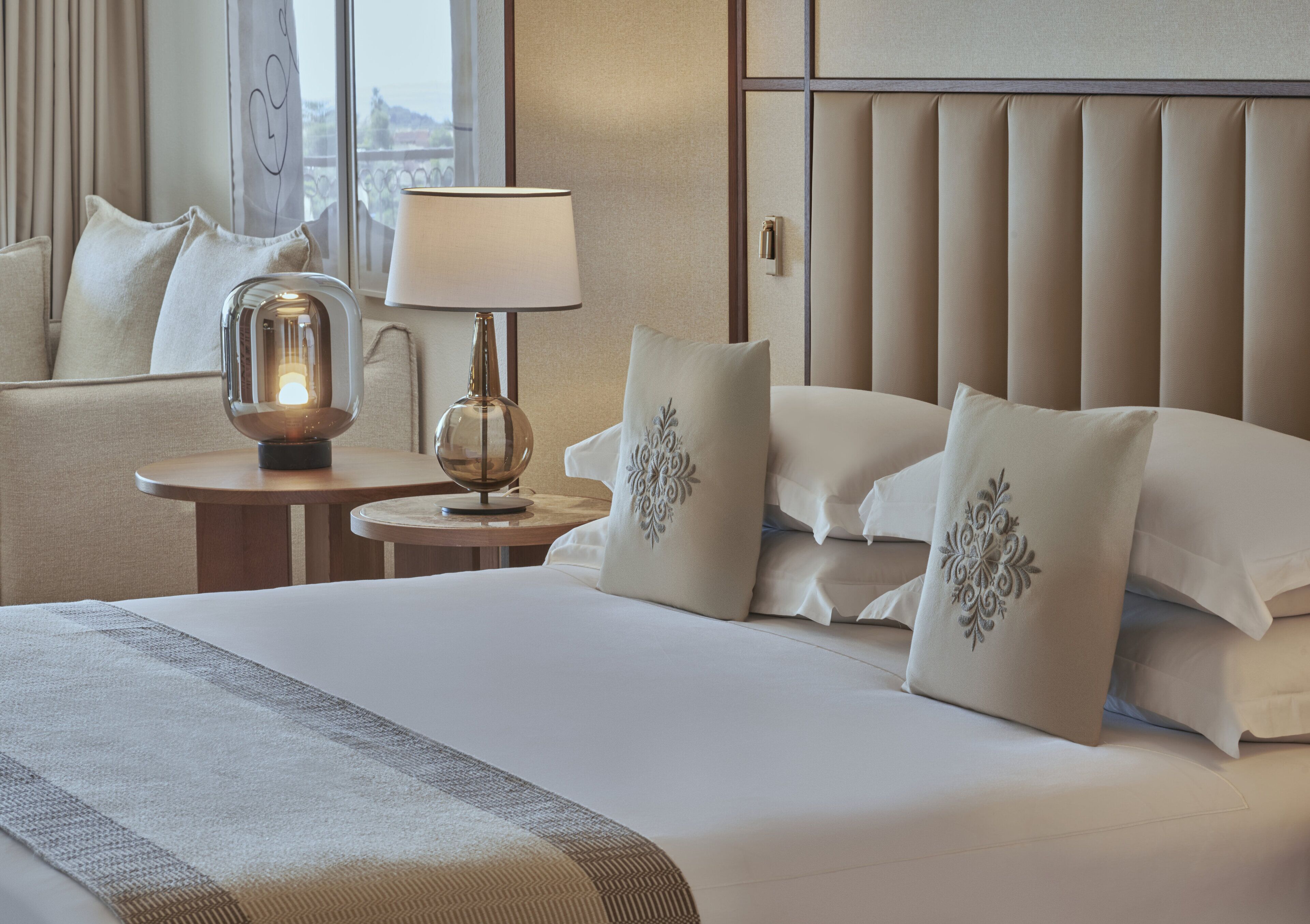 A hotel room with a large bed, a bedside table with a lamp, and a view of the outdoors through a window.