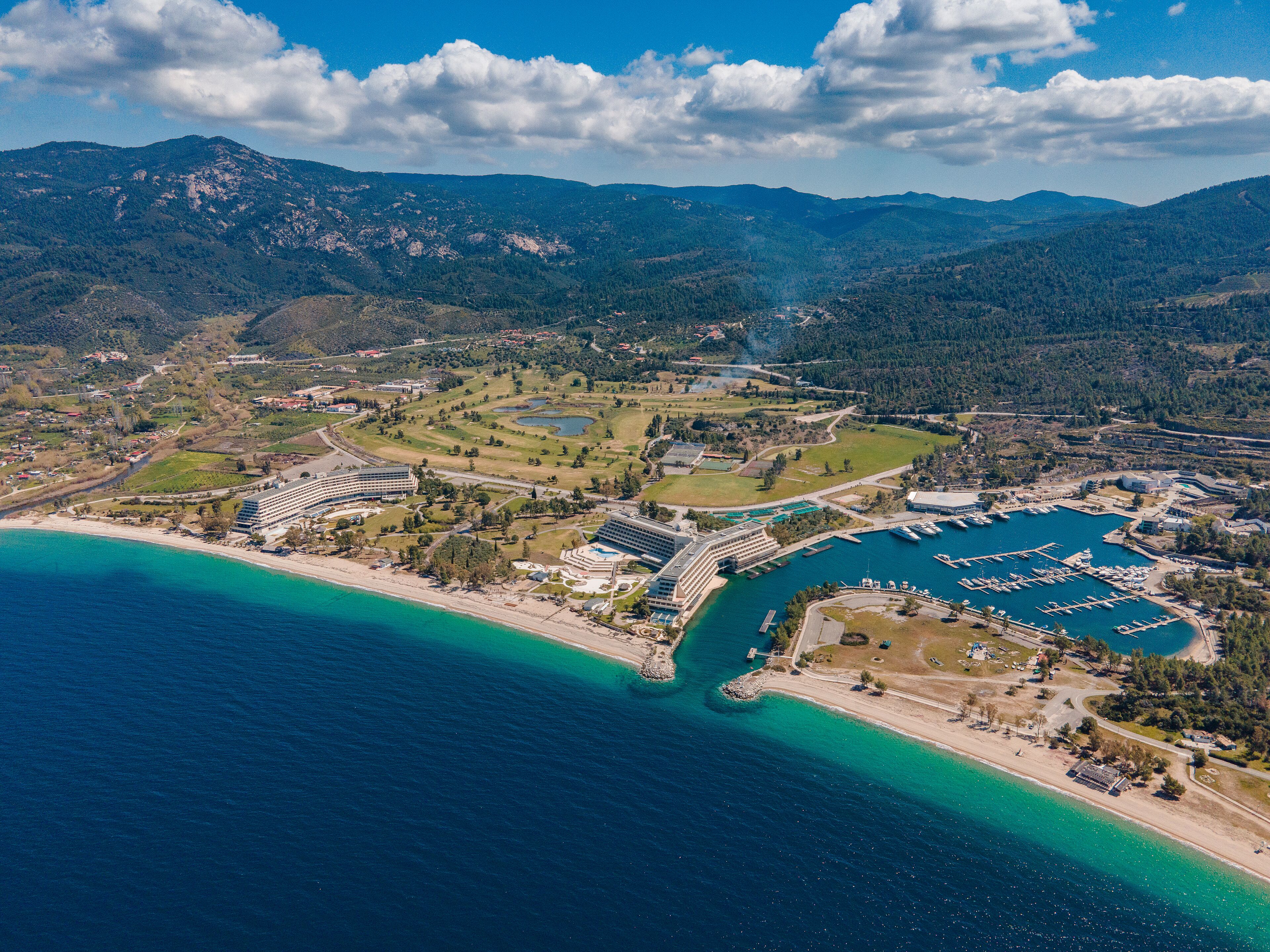 An aerial view of a coastal resort with a marina, beachfront, and mountainous backdrop.
