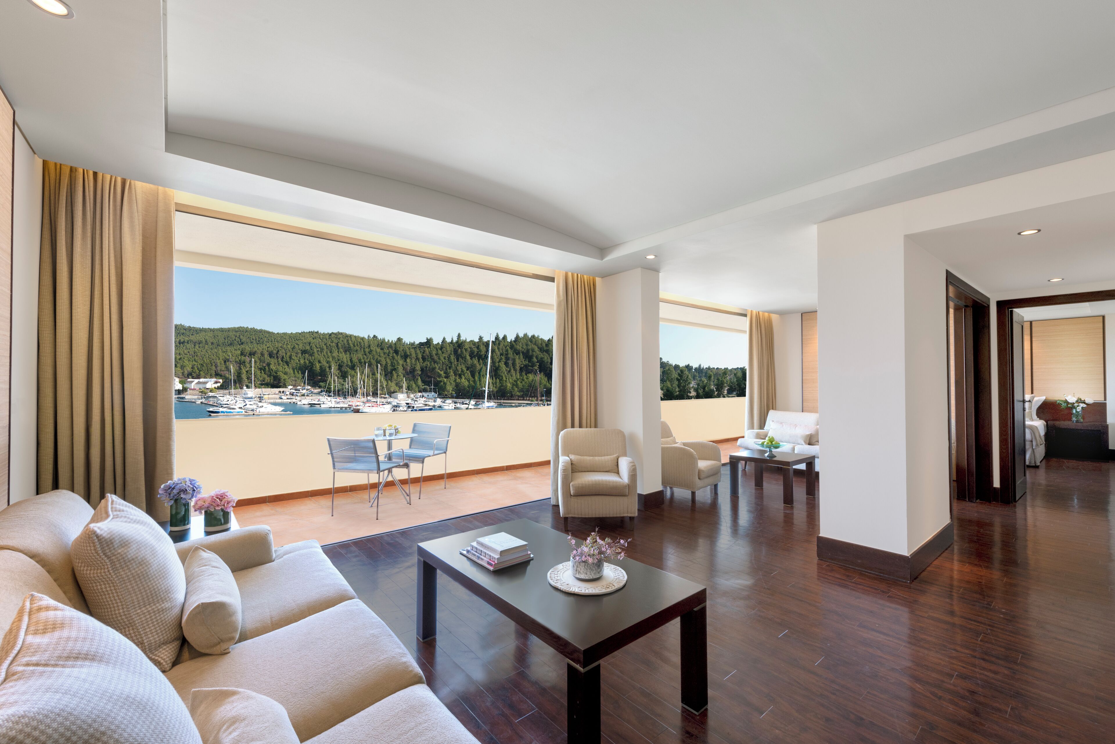 Ein großzügiges Wohnzimmer mit einem großen Fenster mit Blick auf eine Marina und bewaldete Hügel. Es gibt ein Sofa, einen Couchtisch und eine Sitzecke mit Sesseln.