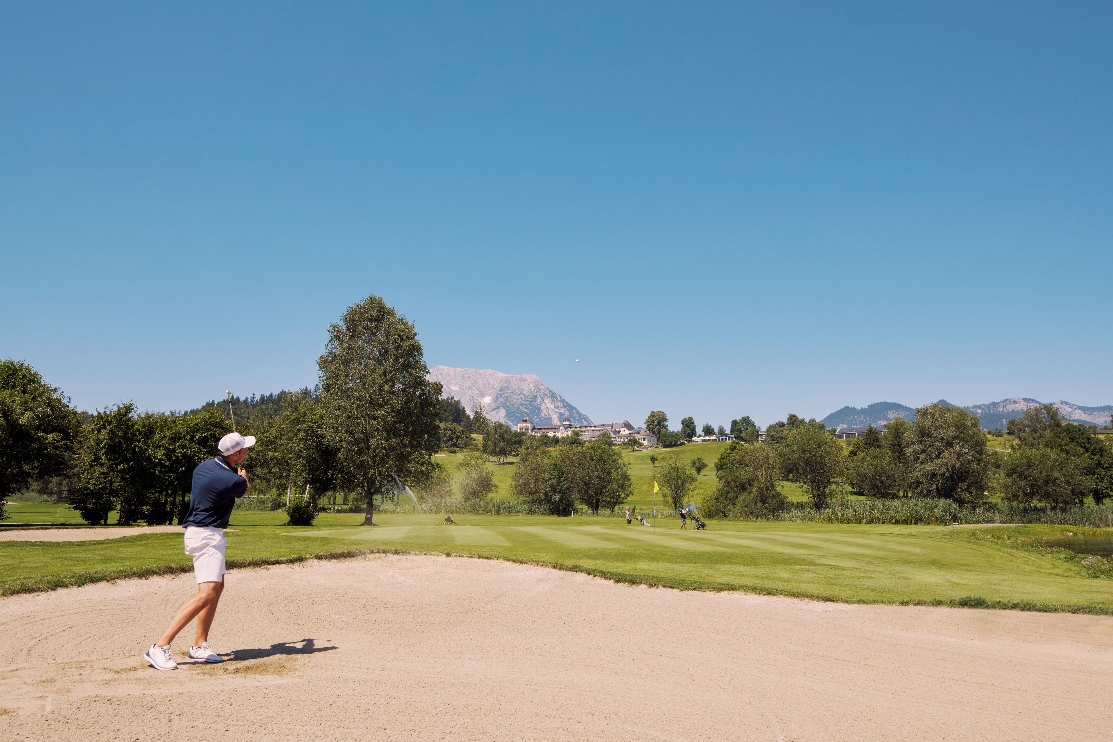 A golf course with a clear sky, a golfer in mid-swing, and a mountainous backdrop.