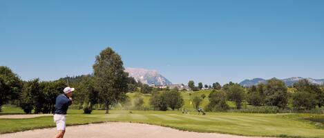 A golf course with a clear sky, a golfer in mid-swing, and a mountainous backdrop.