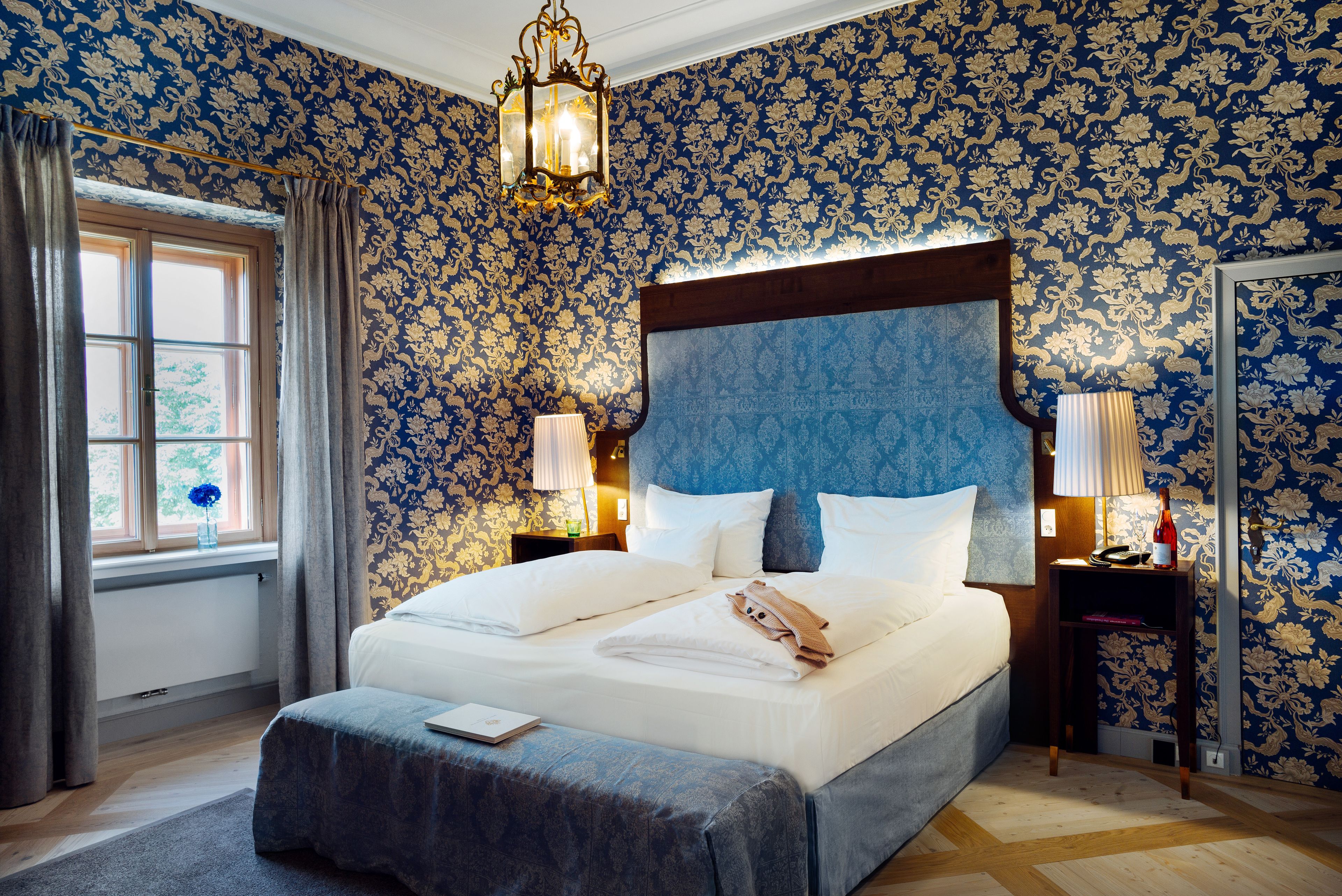 Una cama grande con cabecera de madera, dos mesitas de noche con lámparas, una ventana con cortinas y un estampado floral en el papel tapiz.