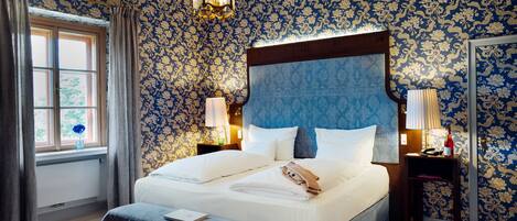 Una cama grande con cabecera de madera, dos mesitas de noche con lámparas, una ventana con cortinas y un estampado floral en el papel tapiz.