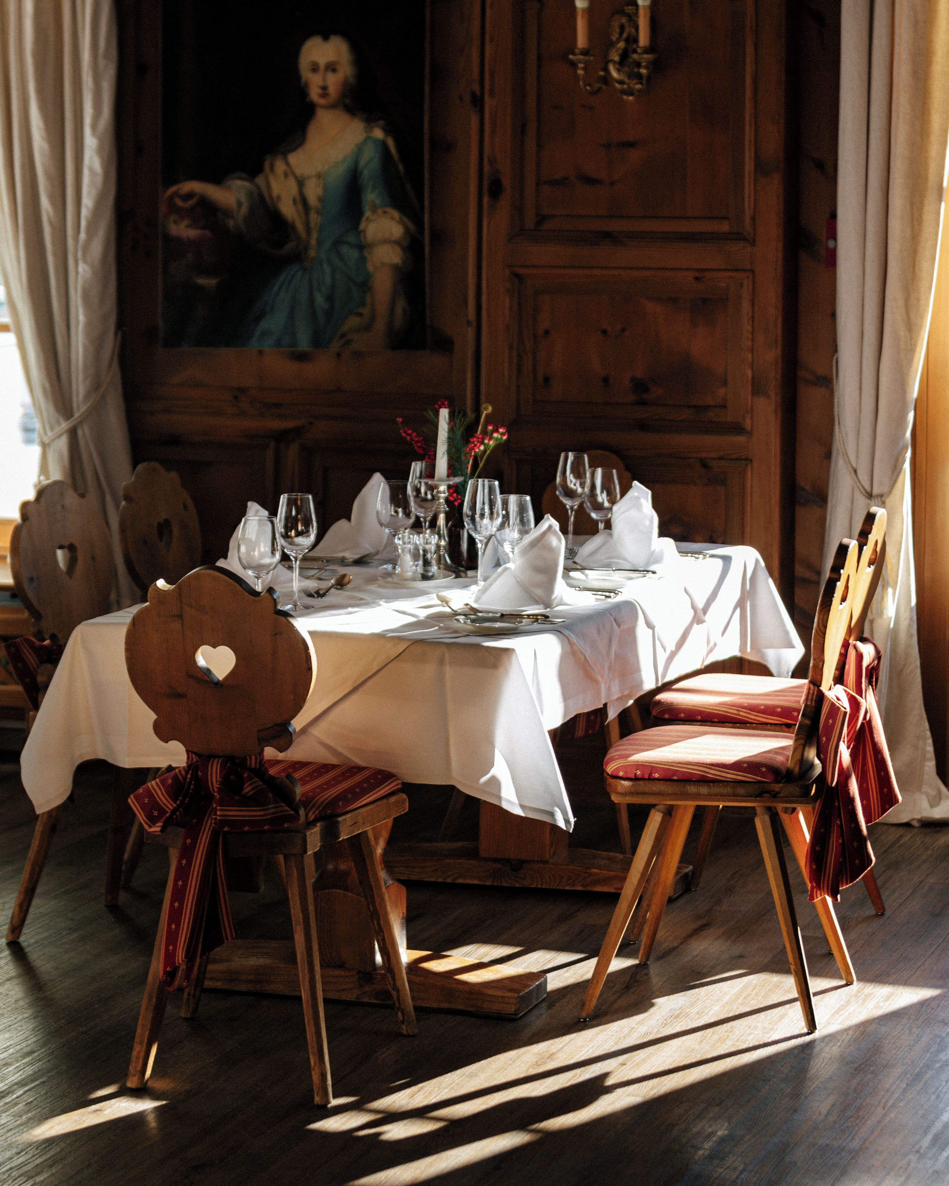 A dining table set for four with a white tablecloth, wooden chairs, and a painting on the wall.