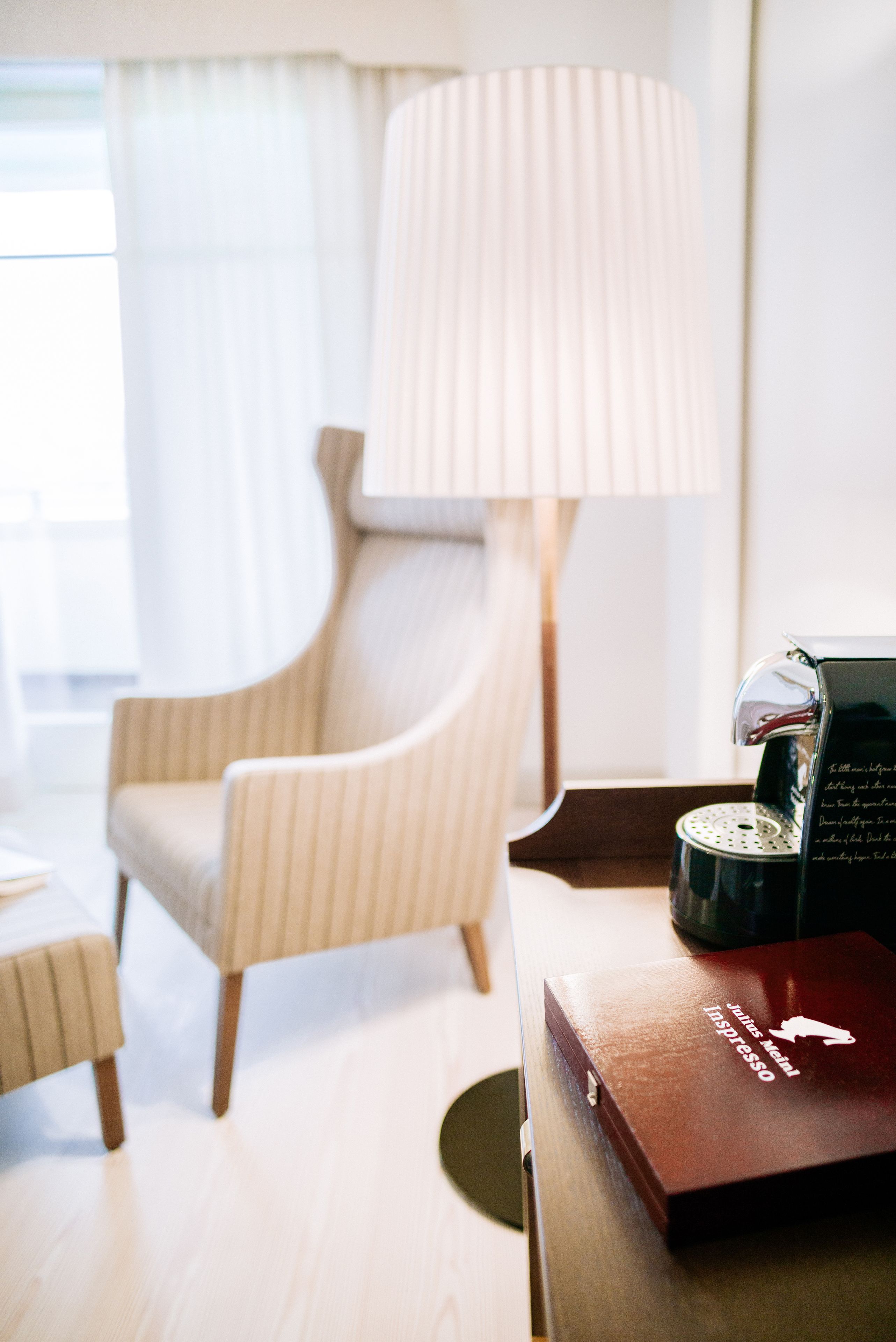 Habitación de hotel con un sillón a rayas, una lámpara y una cafetera sobre una mesa de madera.