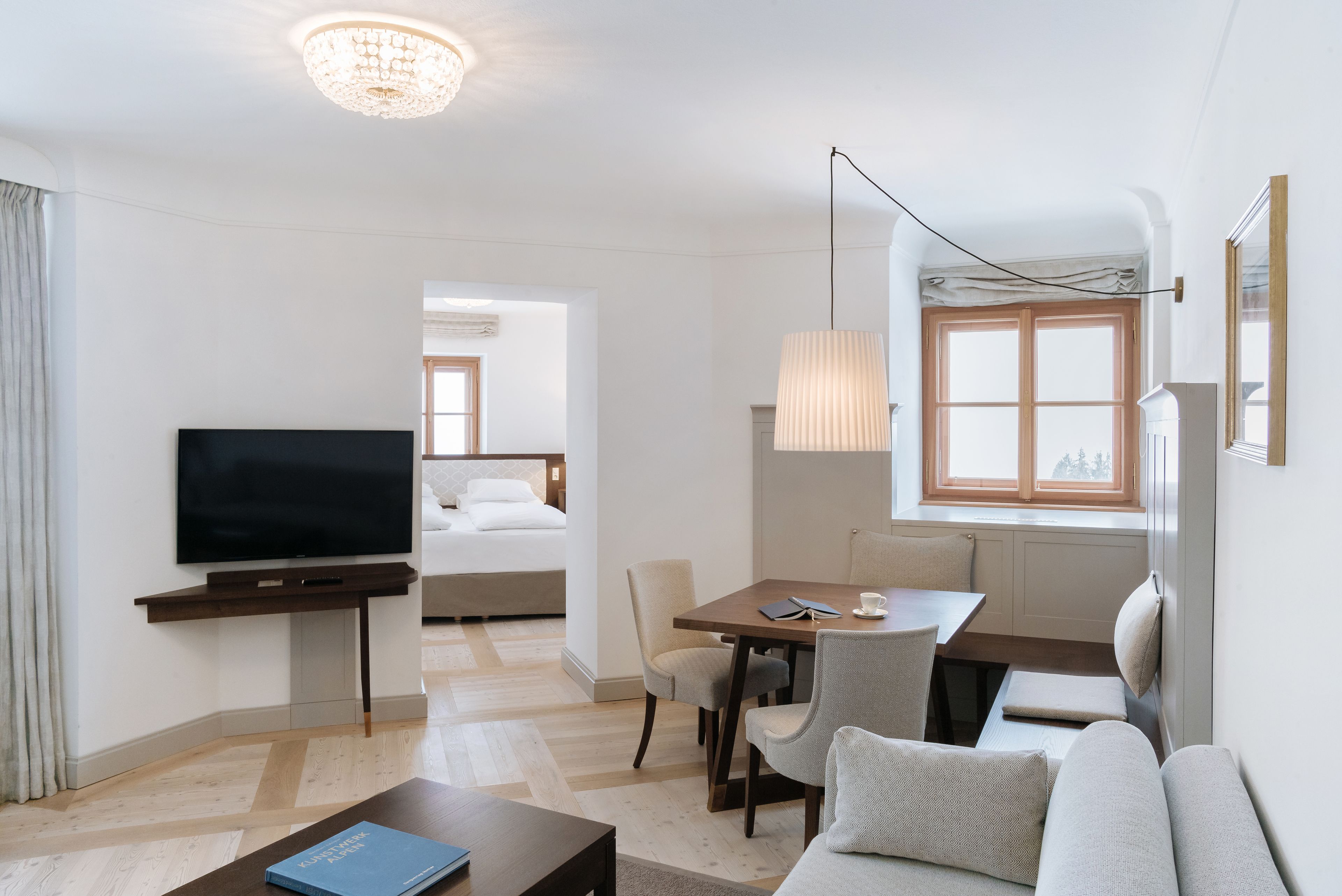 Habitació d'hotel moderna amb televisió de pantalla plana, zona de dinar amb taula i cadires, sofà i finestra gran amb llum natural.