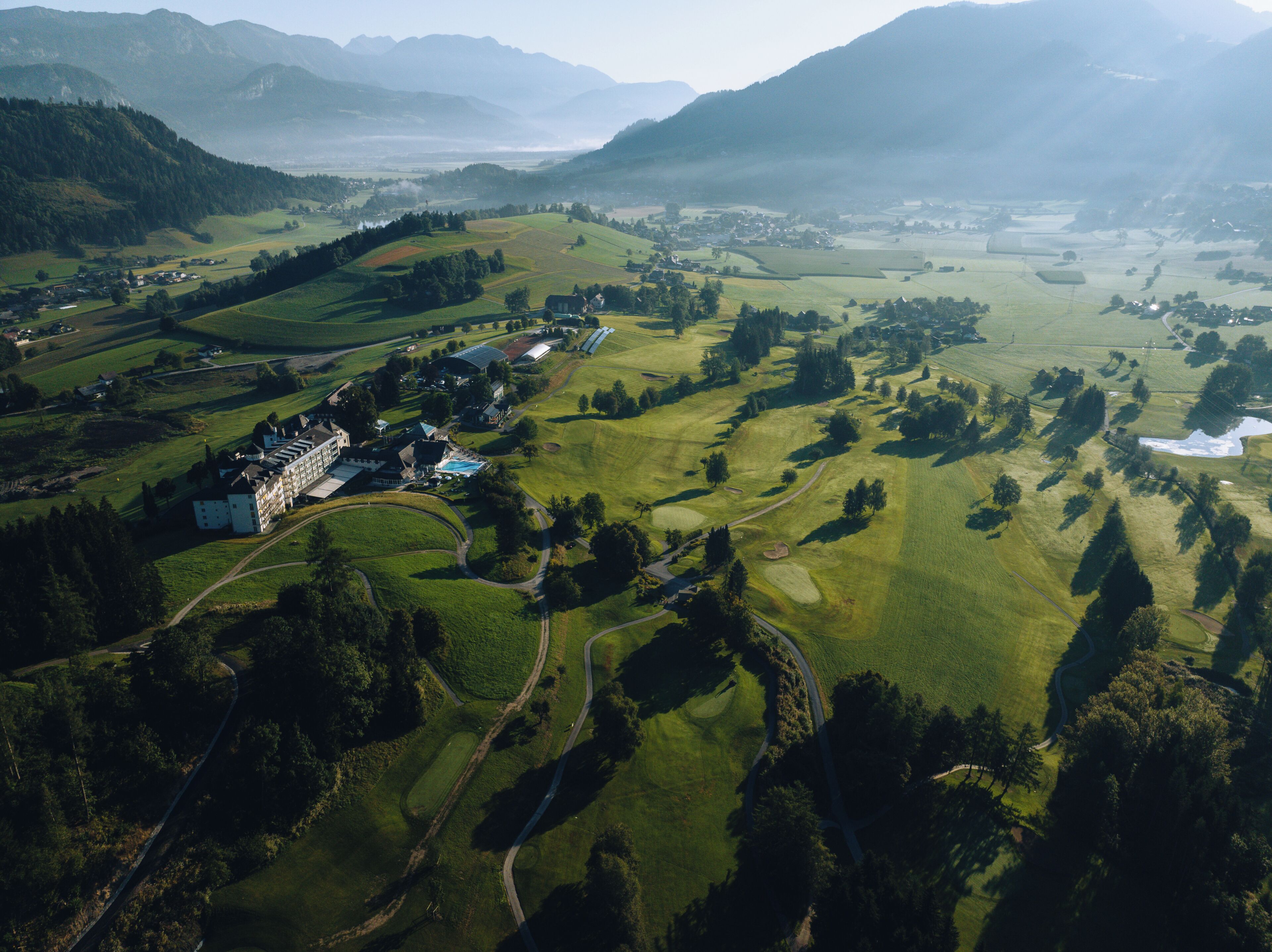 An aerial view of a golf course with a clubhouse, surrounded by rolling hills and mountains.