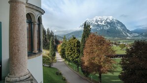 A building with a classical architectural style, featuring a prominent column and arched windows. In the background, there is a snow-capped mountain and a green landscape with trees and a pathway leading towards a village.