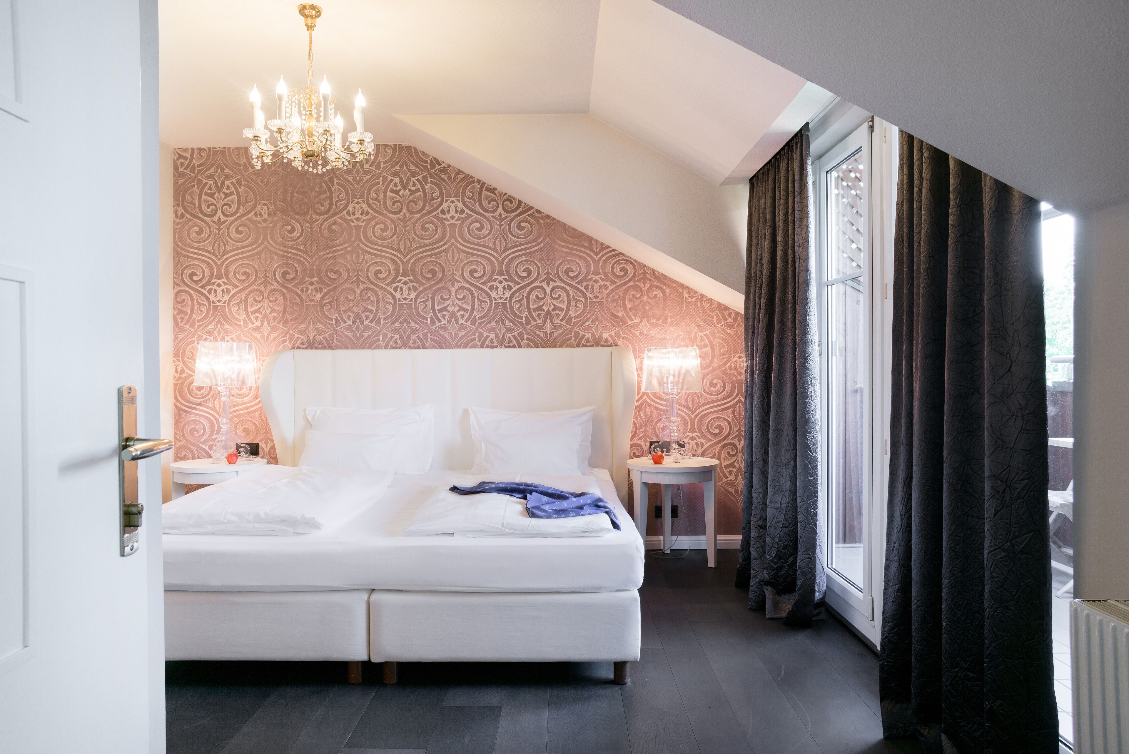 A bedroom with a large bed, two bedside tables, a chandelier, patterned wallpaper, and a window with curtains.
