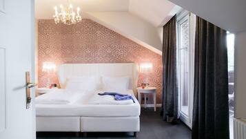 A bedroom with a large bed, two bedside tables, a chandelier, patterned wallpaper, and a window with curtains.