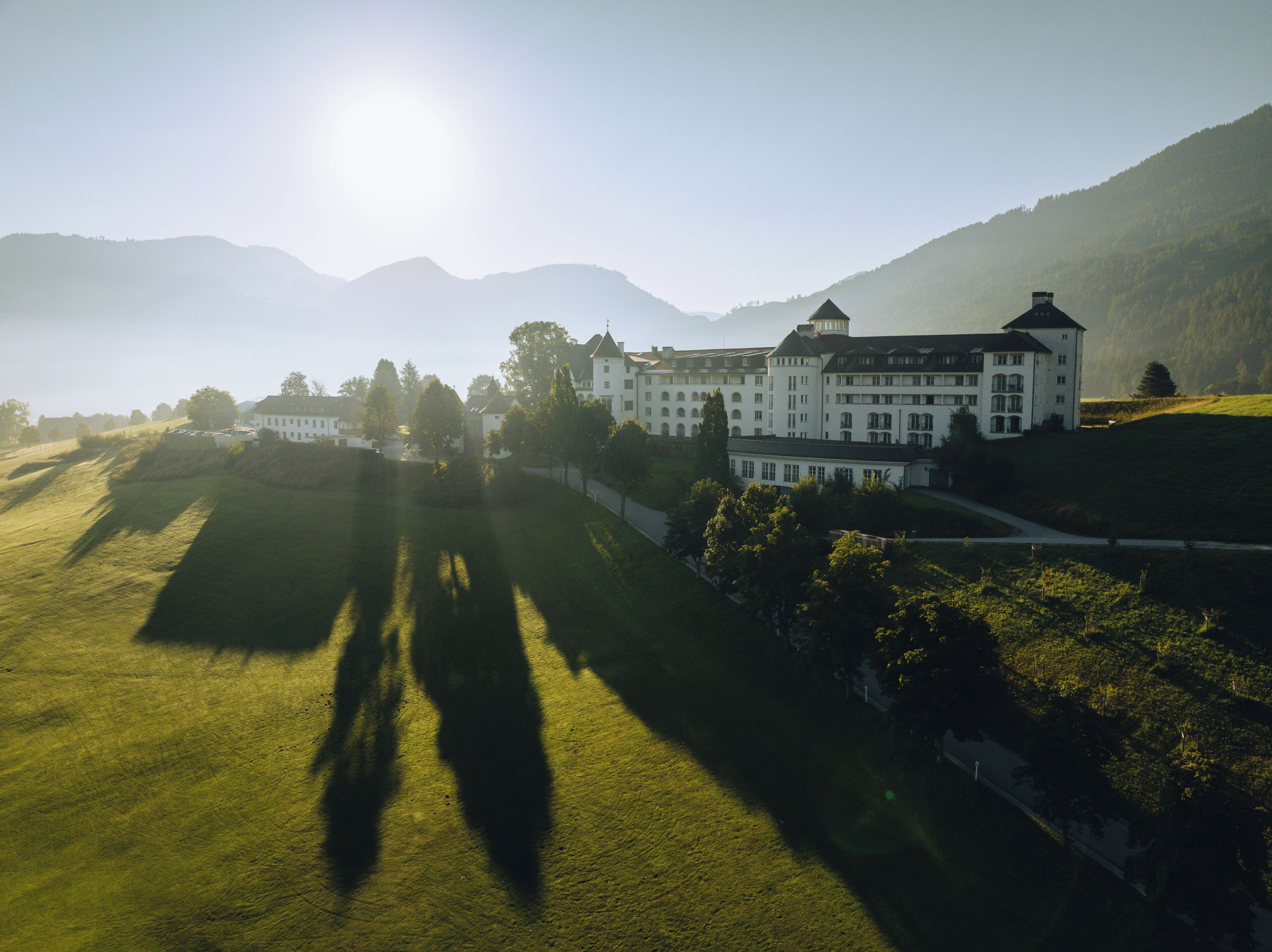 A large, white building with multiple windows and a prominent tower, surrounded by green fields and mountains.