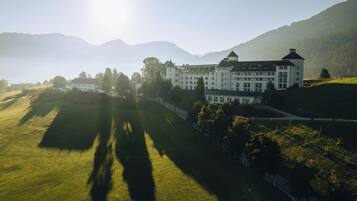 A large, white building with multiple windows and a prominent tower, surrounded by green fields and mountains.