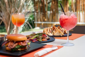 Un pasto con hamburger, verdure grigliate e un panino, accompagnato da due bevande, una arancione e una rosa, su un tavolo all'aperto.